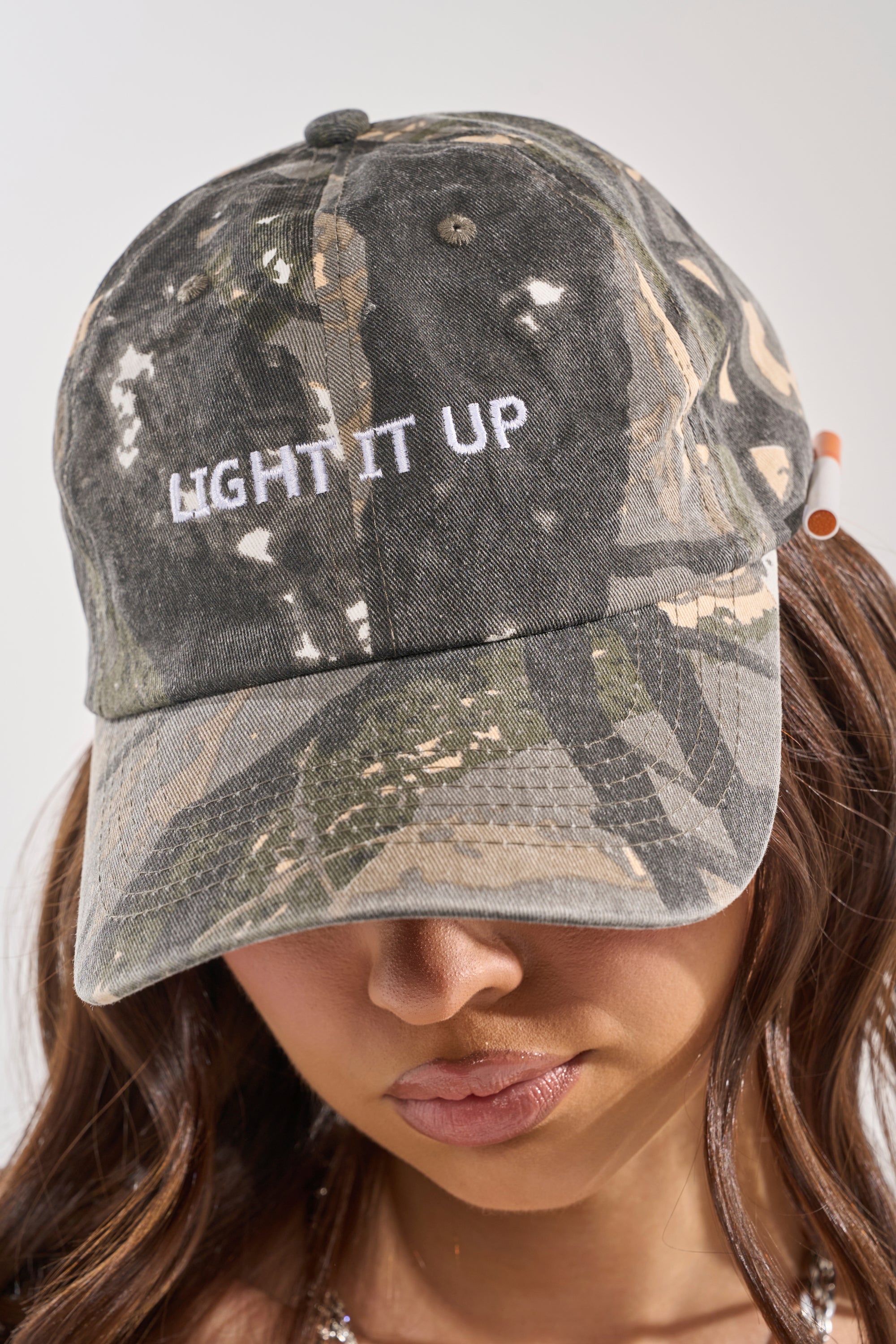 A person wears the LIGHT IT UP FAUX CIG HAT—a camo baseball cap with "LIGHT IT UP" in white embroidery—their face partially visible from the nose down. 2