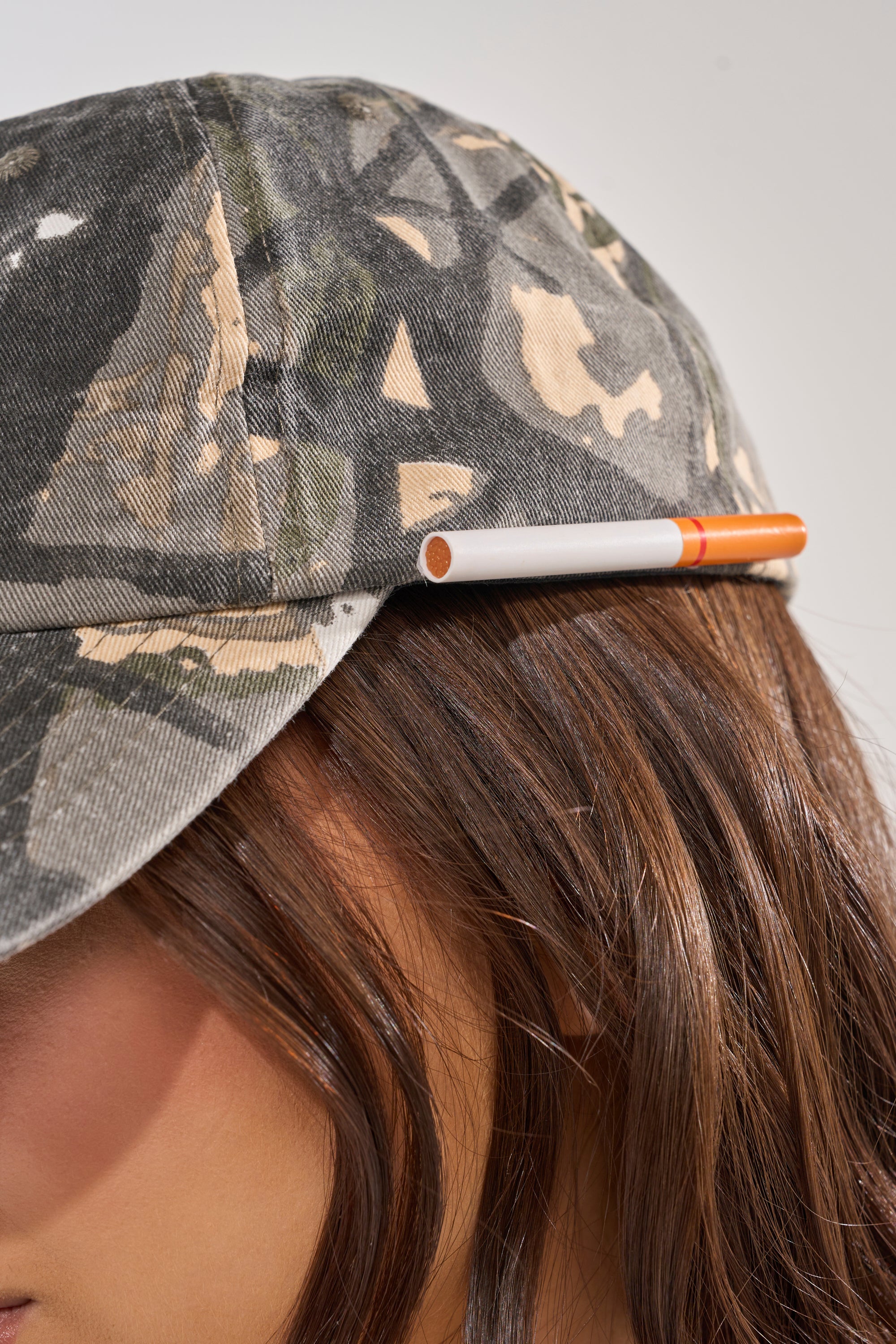A close-up of a person wearing the LIGHT IT UP FAUX CIG HAT, a camo print baseball cap featuring a faux cigarette above the ear. Only part of their face and brown hair are visible, highlighting casual street style. 3