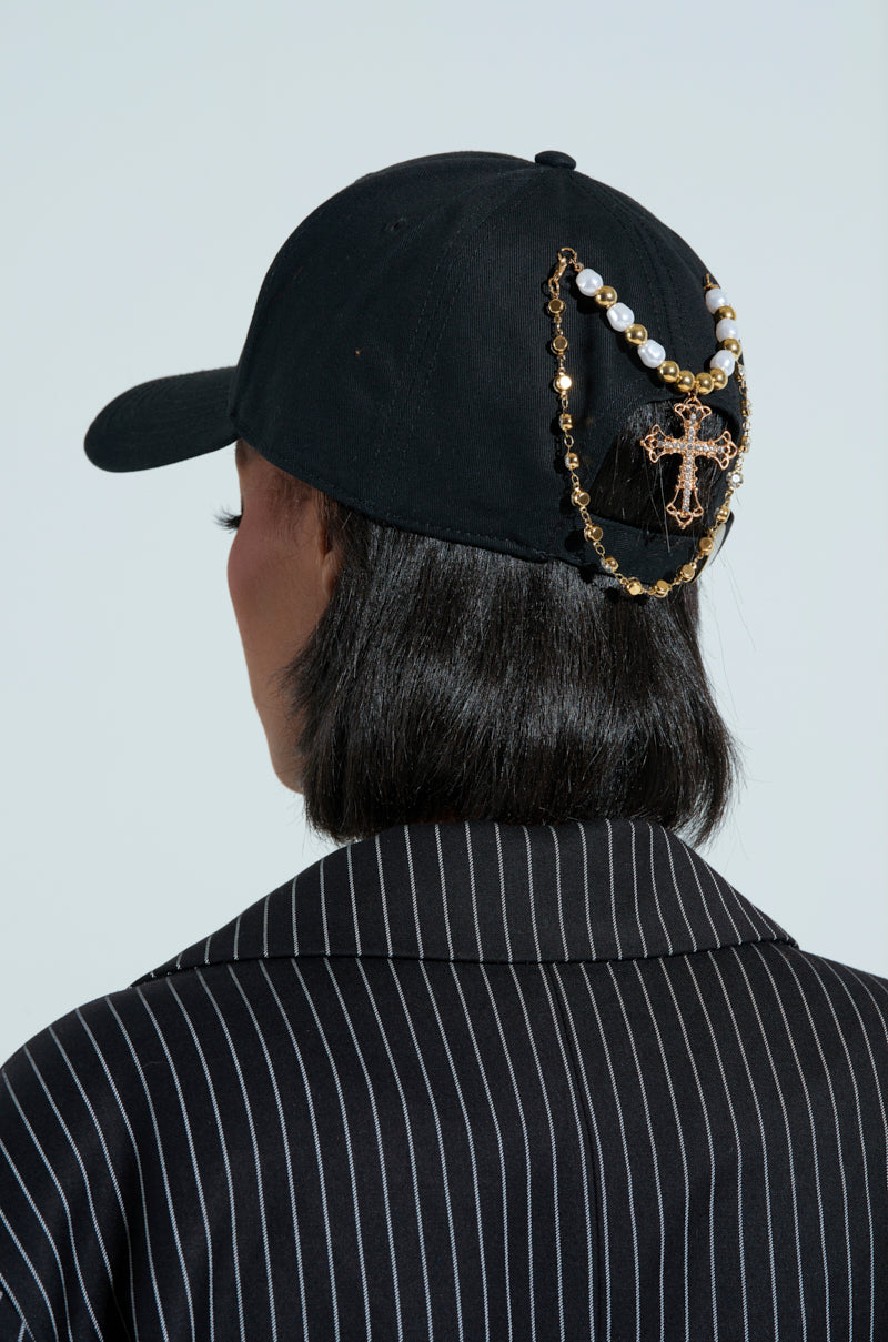 A person with short black hair wears the PRAY FOR YOU ROSARY HAT, featuring faux gold chains, pearl beads, and a decorative cross on the back. They pair it with a black pinstripe jacket against a plain light-colored background.