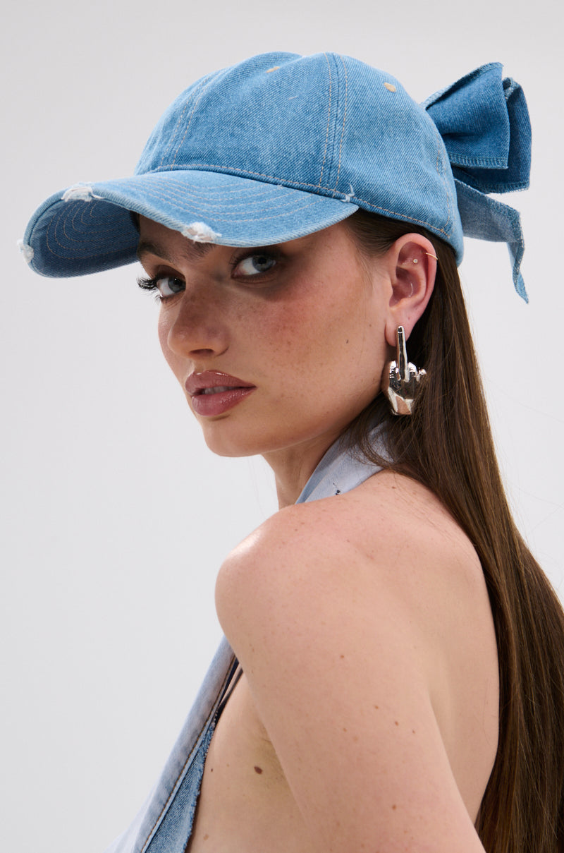 A woman models the DENIM DENIM DENIM HAT—a distressed light blue denim baseball cap with an adjustable strap and large back bow—styled with silver hoop earrings and a denim halter top, glancing over her shoulder against a plain backdrop.