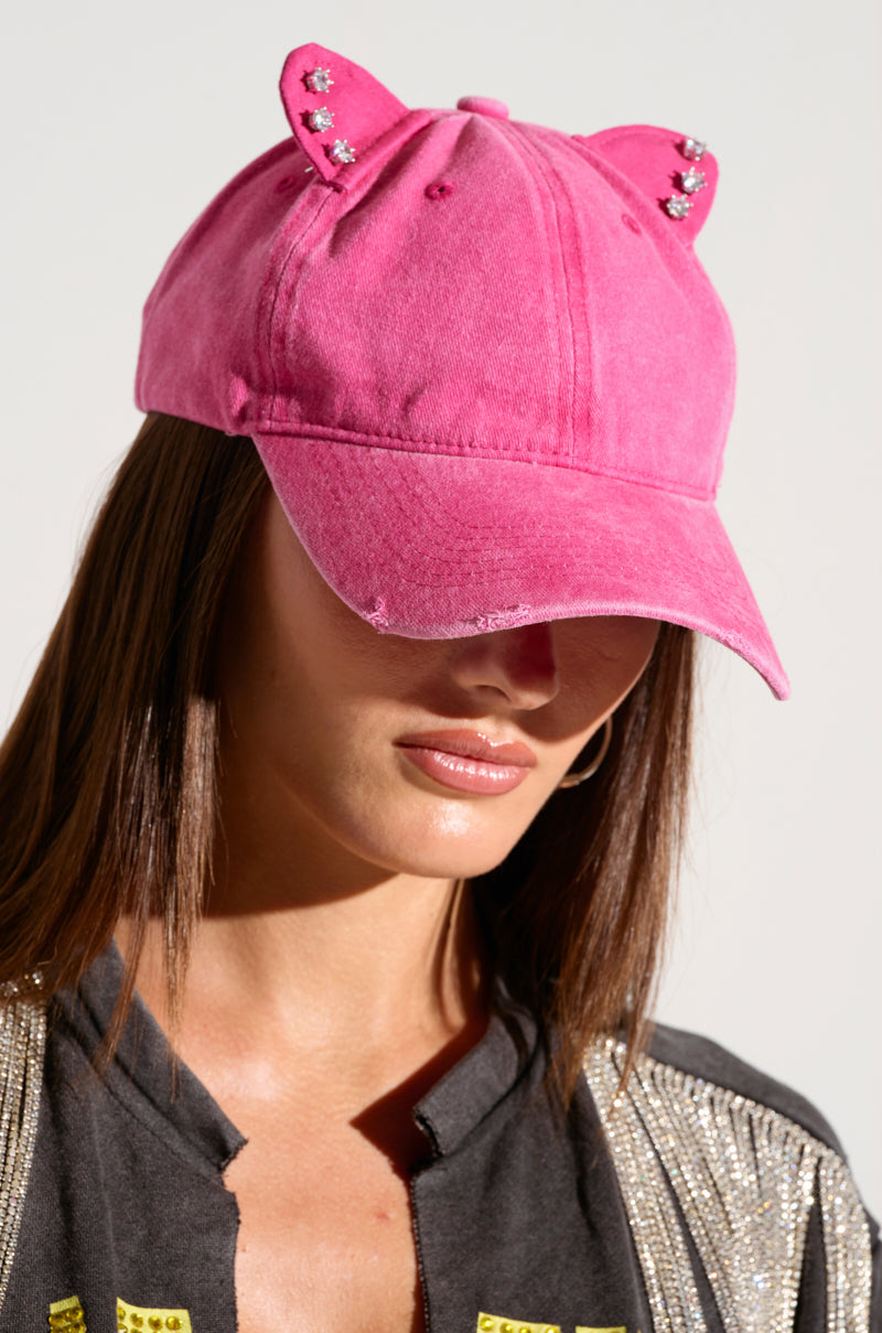 A person wearing a hot pink TOO COI SNAPBACK embellished with cat ears and small rhinestones. The individual has long brown hair and is dressed in a black outfit with gold accents. The person's face is partially visible, and they are looking slightly downward.