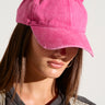 A person wearing a hot pink TOO COI SNAPBACK embellished with cat ears and small rhinestones. The individual has long brown hair and is dressed in a black outfit with gold accents. The person's face is partially visible, and they are looking slightly downward.