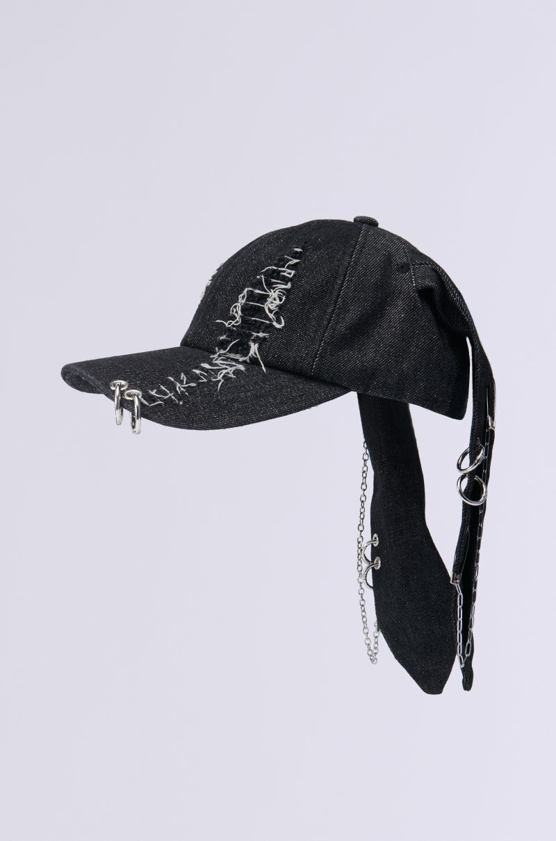 The AERIAL VIEWS HAT is a black denim baseball cap embellished with graffiti-style writing along the brim. It features metallic silver accessories such as attached chains and decorative rings, and includes additional fabric draped like a scarf from the back, set against a plain, light background. 4