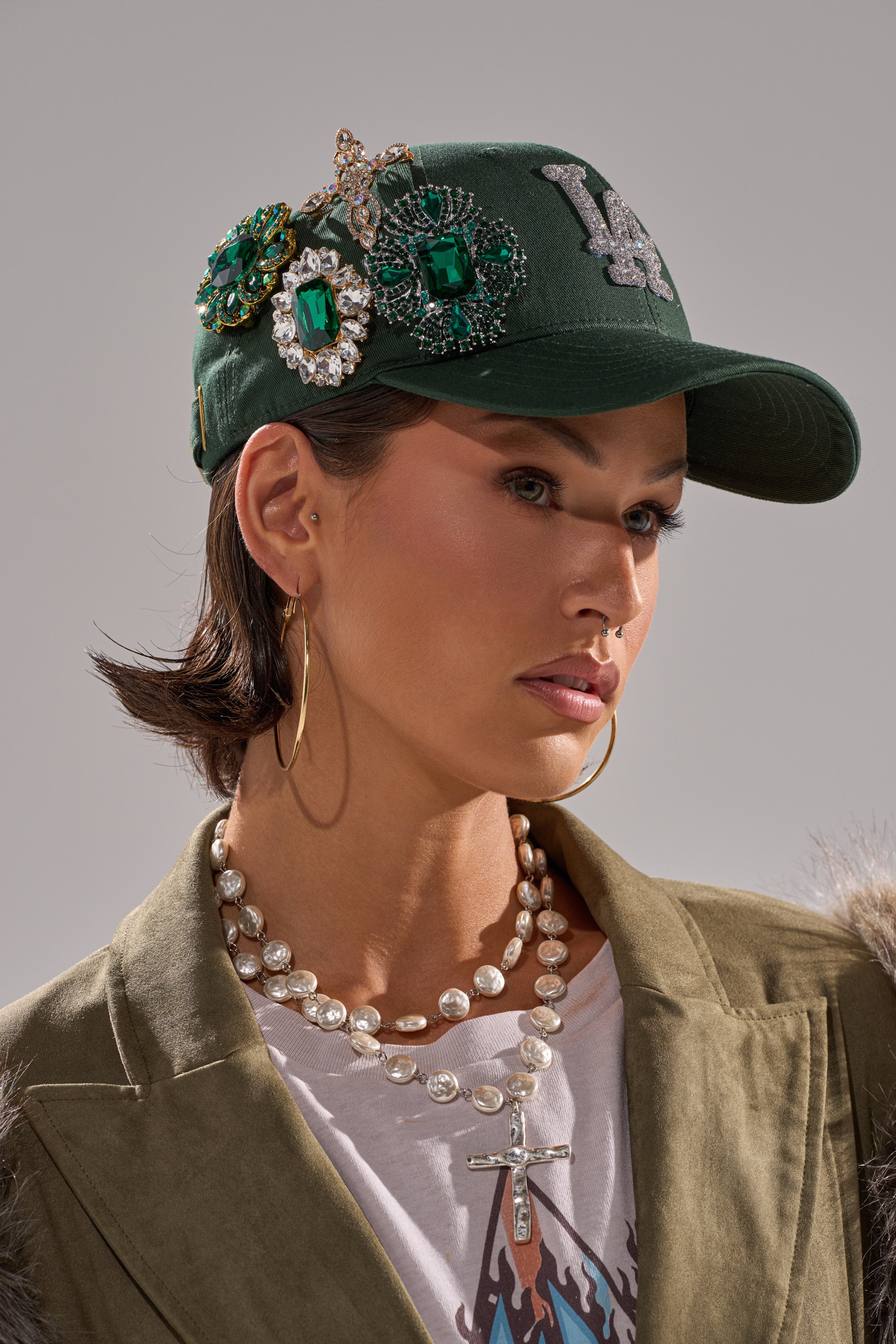 A woman poses against a neutral background wearing an EMMIE BROOCH HAT in green, embellished with jeweled brooches, along with gold hoop earrings, layered pearl necklaces, a suede jacket, and a white T-shirt.