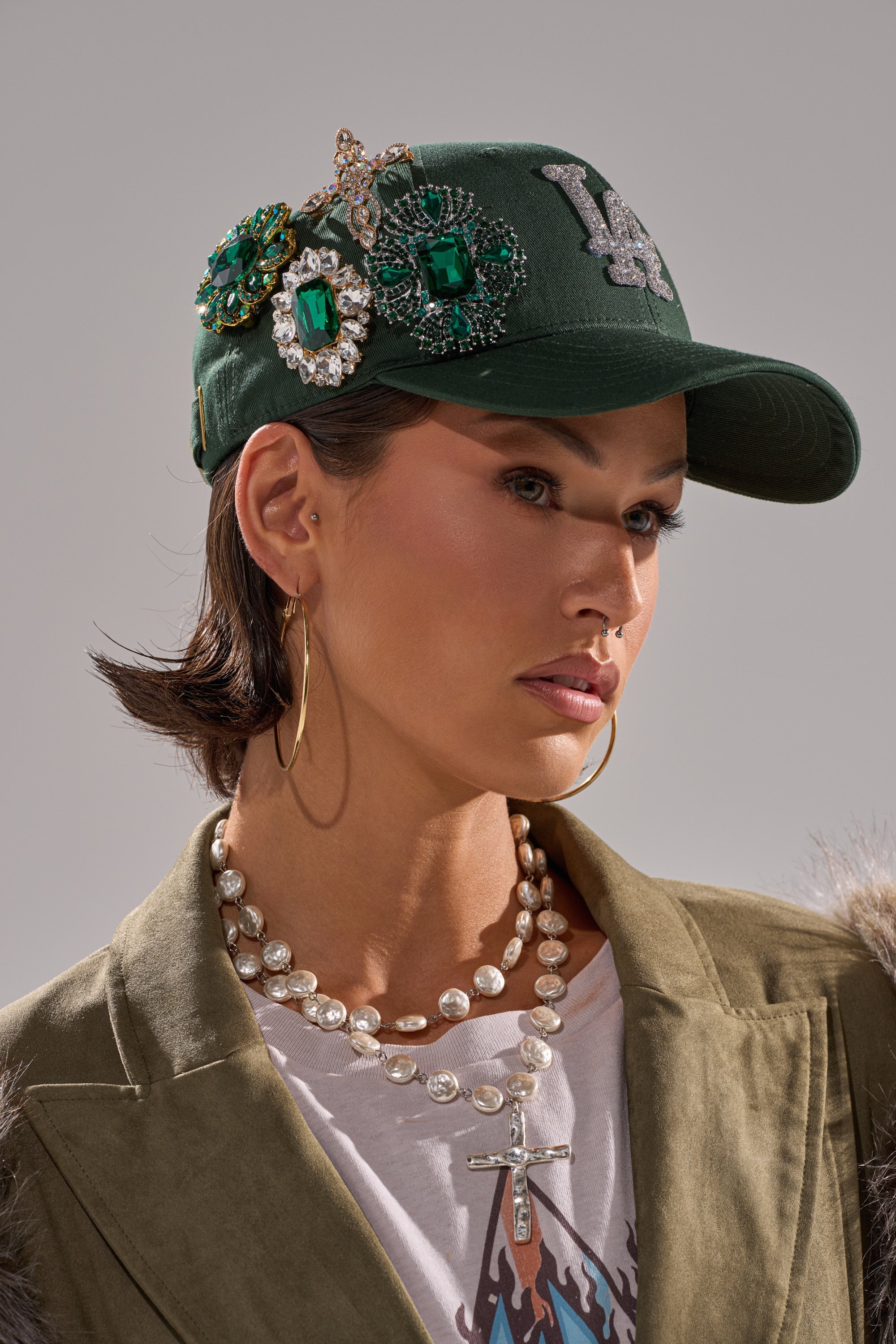 A woman poses against a neutral background wearing an EMMIE BROOCH HAT in green, embellished with jeweled brooches, along with gold hoop earrings, layered pearl necklaces, a suede jacket, and a white T-shirt. 0