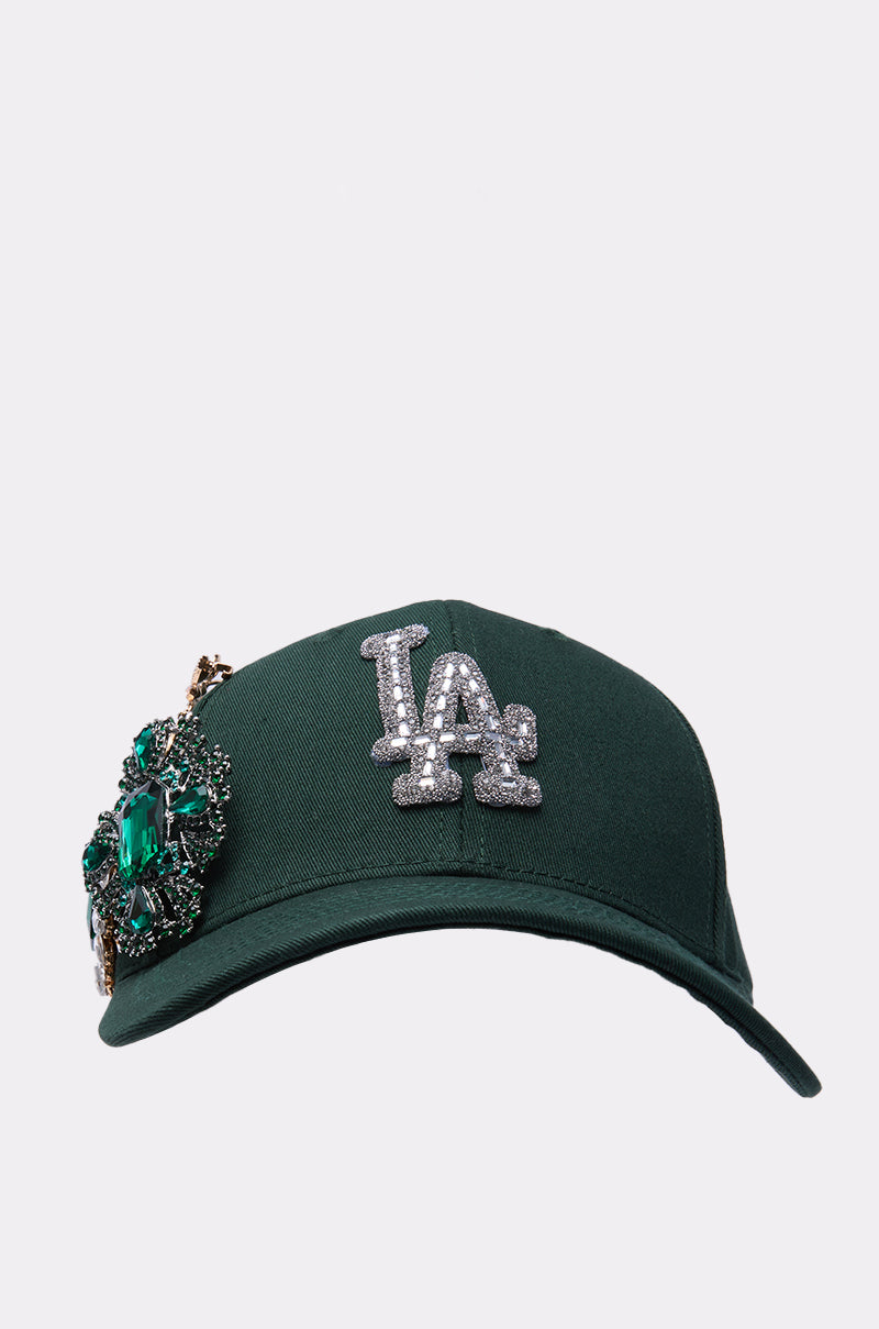 The EMMIE BROOCH HAT is a dark green rhinestone baseball cap with a jeweled LA logo and a large green gemstone brooch on the left front panel, shown against a plain white background. 2