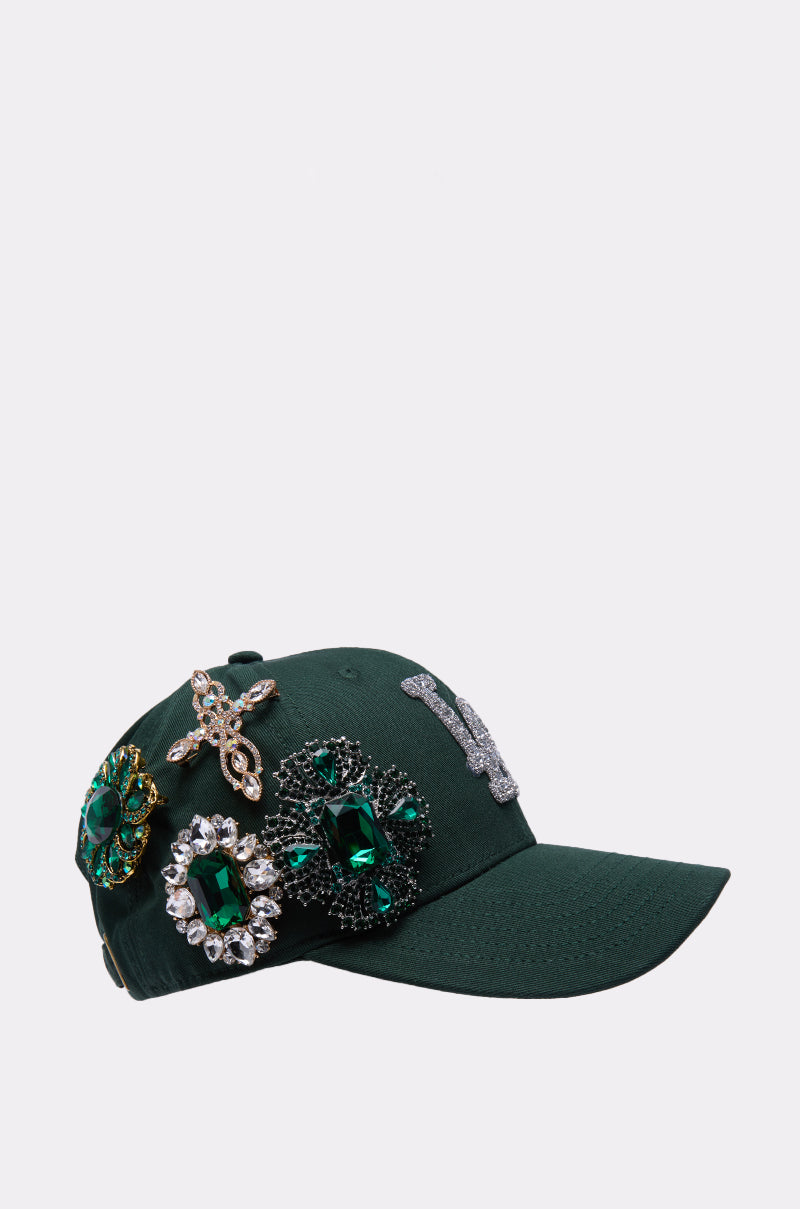 The EMMIE BROOCH HAT is a dark green rhinestone baseball cap adorned with bold green and gold jeweled brooches and a glittering silver "W" on the front, shown against a simple light-colored background.