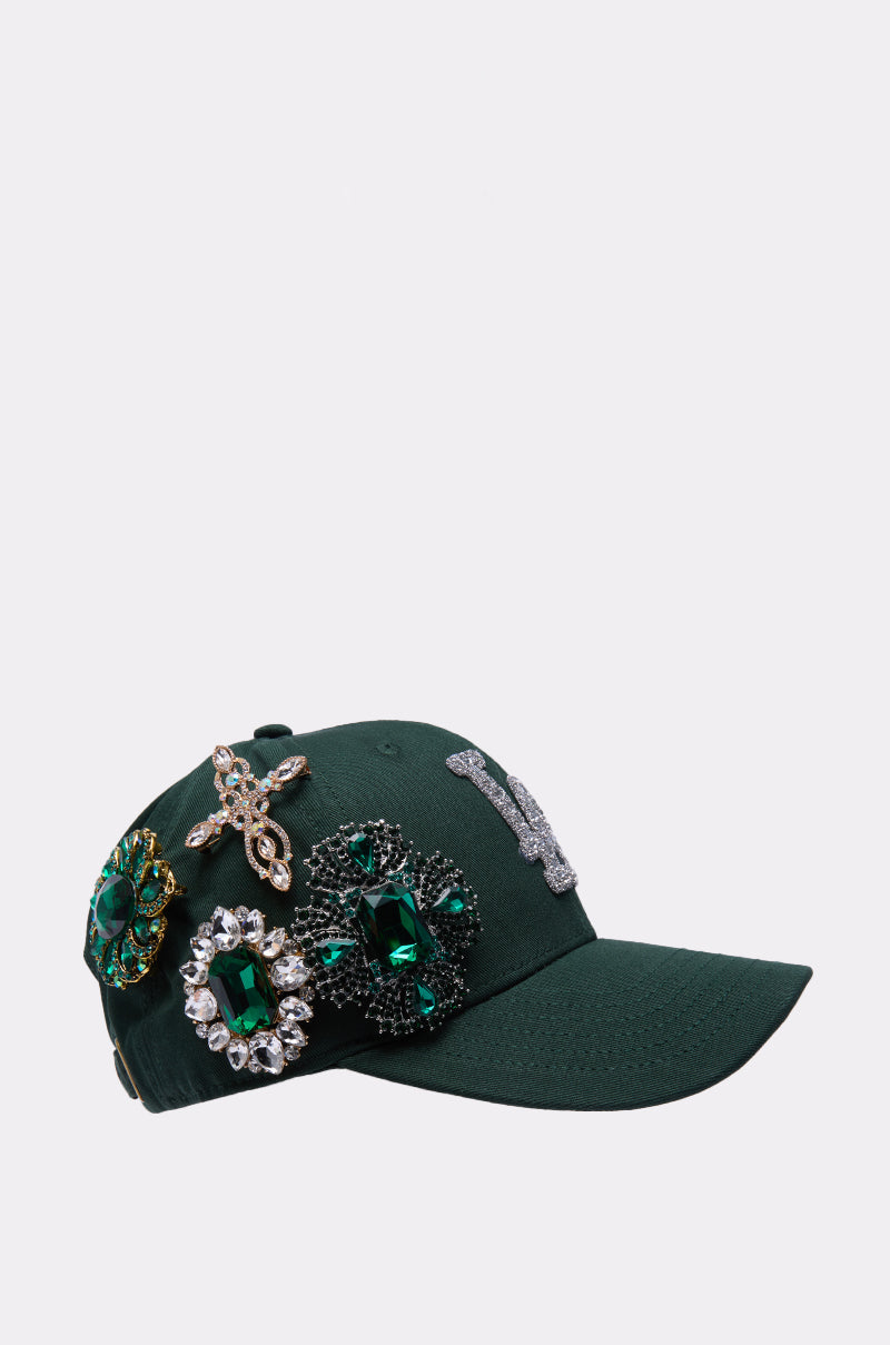 The EMMIE BROOCH HAT is a dark green rhinestone baseball cap adorned with bold green and gold jeweled brooches and a glittering silver "W" on the front, shown against a simple light-colored background. 3