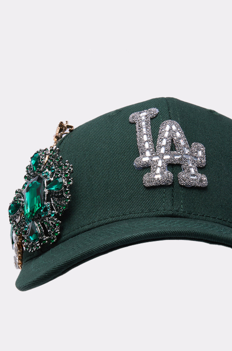 The EMMIE BROOCH HAT is a dark green rhinestone baseball cap with a sparkling silver "LA" logo and an ornate green and silver jeweled brooch on the front left side.