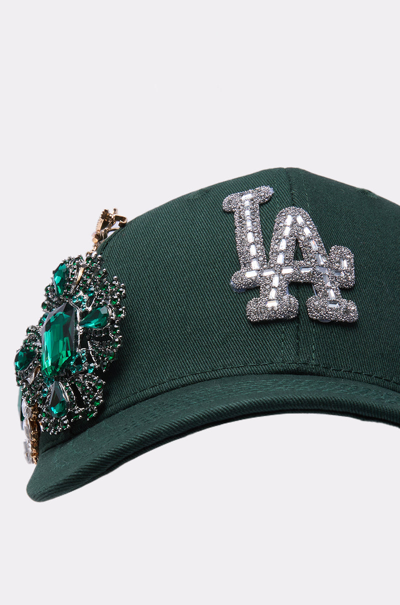 The EMMIE BROOCH HAT is a dark green rhinestone baseball cap with a sparkling silver "LA" logo and an ornate green and silver jeweled brooch on the front left side. 4