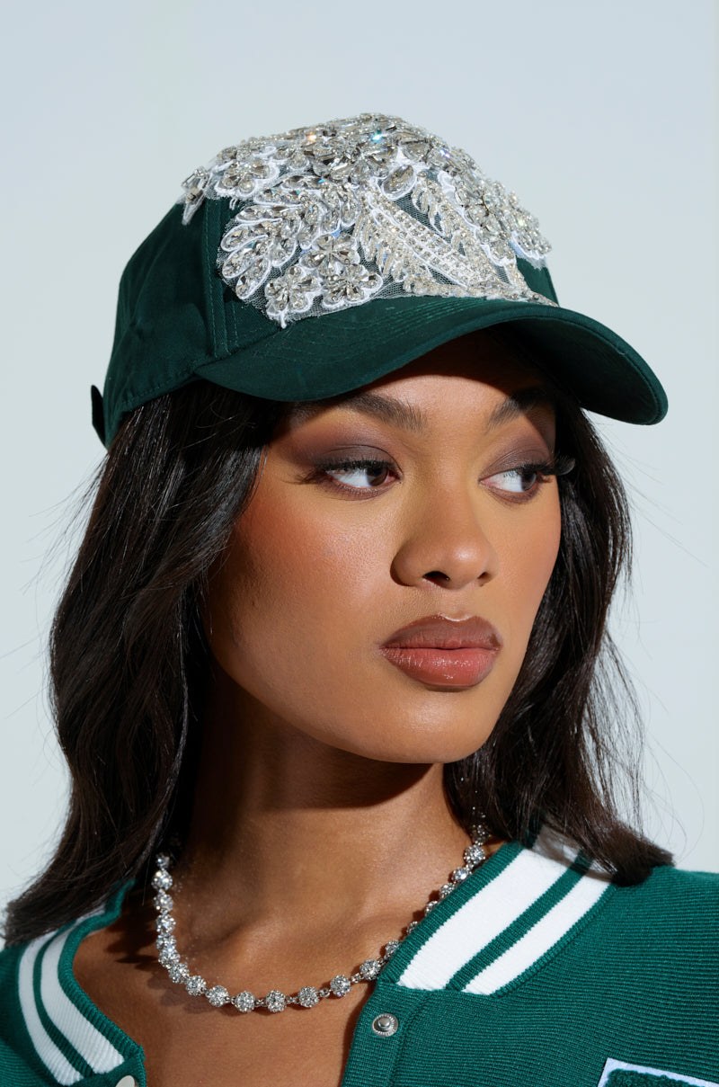 A woman with a neutral expression and long, dark hair wears the FOREVER EXTRA APPLIQUE HAT from the AKIRA Label—a dark green baseball cap adorned with rhinestones. She pairs it with a green jacket featuring white-striped collar and a sparkly necklace. The background is plain. 0