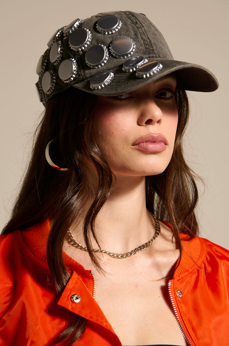 A woman with long brown hair poses against a neutral background wearing the BOTTLES ON ME HAT from AKIRA Label, featuring bottle cap embellishments, along with a silver hoop earring, chain necklace, orange jacket, and black top.