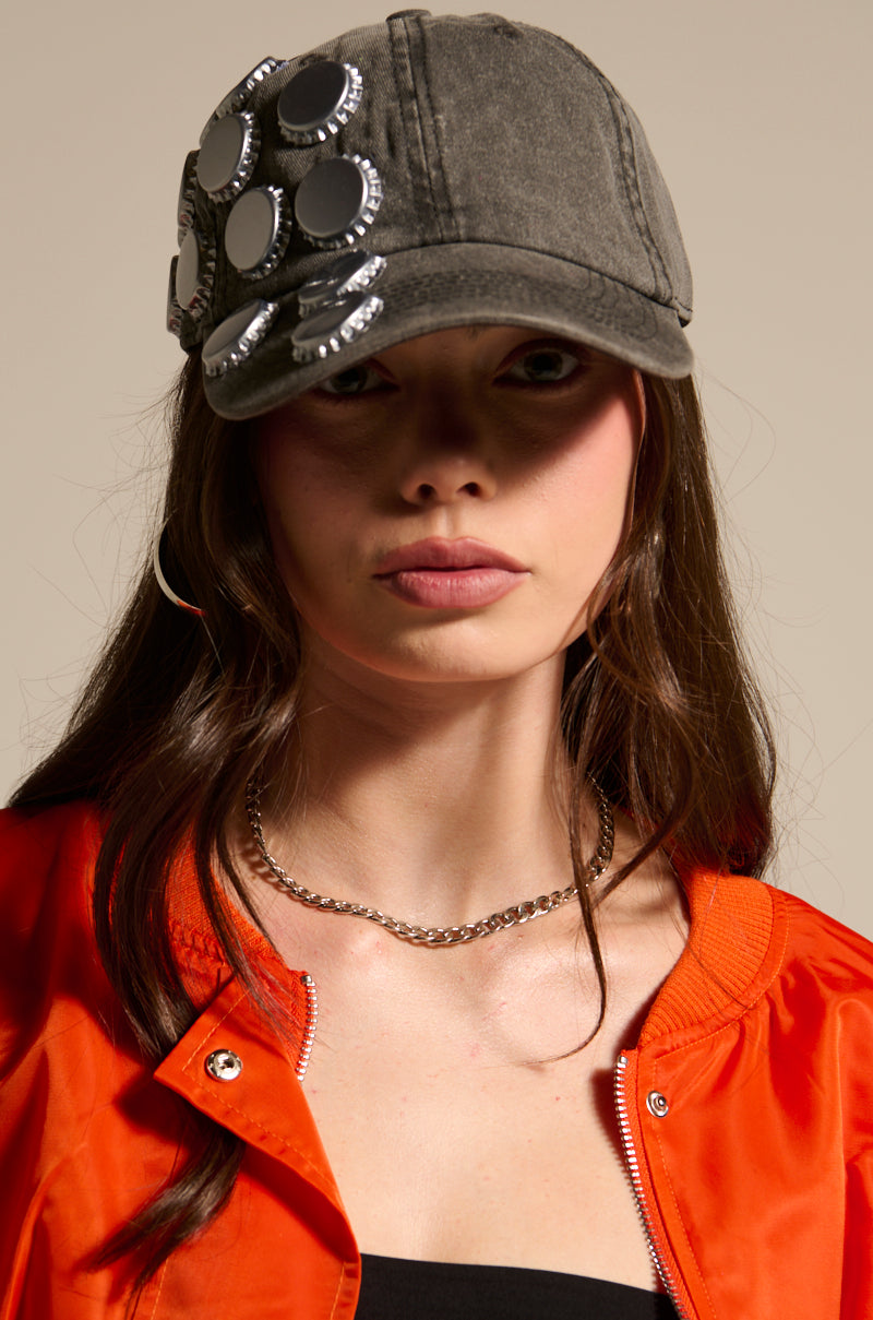 A woman poses against a neutral background wearing the BOTTLES ON ME HAT—a gray baseball cap with round, bottle cap embellishments—paired with a chunky silver chain necklace, large hoop earrings, and an orange jacket over a black top.