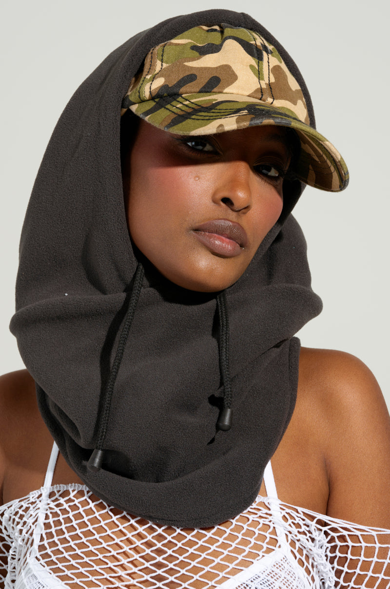 A person wearing the BROOKLYN CAP AND HOOD with a camo pattern and a dark hooded top featuring loose ties. They have a relaxed expression, complemented by a white mesh top, all set against a neutral background.