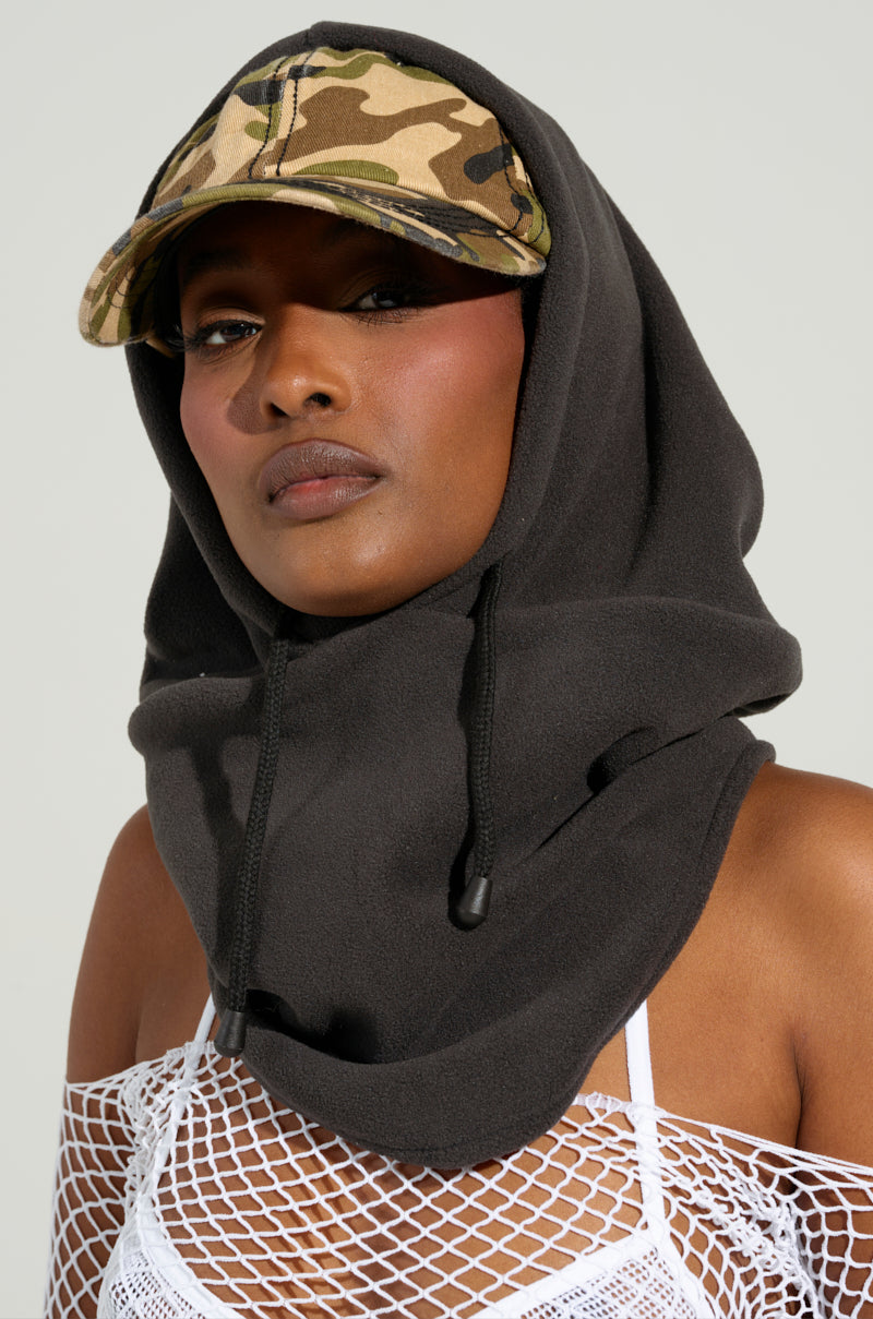 A person wearing a BROOKLYN CAP AND HOOD with a camo pattern layered over a white fishnet top, staring directly at the camera against a plain, light-colored background.