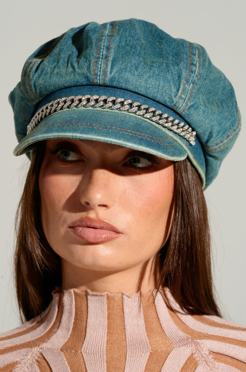 A close-up of a woman wearing the DENIM BLUES BRIM HAT, featuring an intricately designed rhinestone-embellished chain just above the brim. She has long brown hair and is dressed in a pink and beige striped top. Her expression remains neutral against a plain light gray background. 2