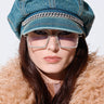 A person with long brown hair is wearing the DENIM BLUES BRIM HAT adorned with a rhinestone embellished chain, large square rhinestone glasses, and a fuzzy tan-colored jacket. The background is plain white, and the person's expression is neutral.