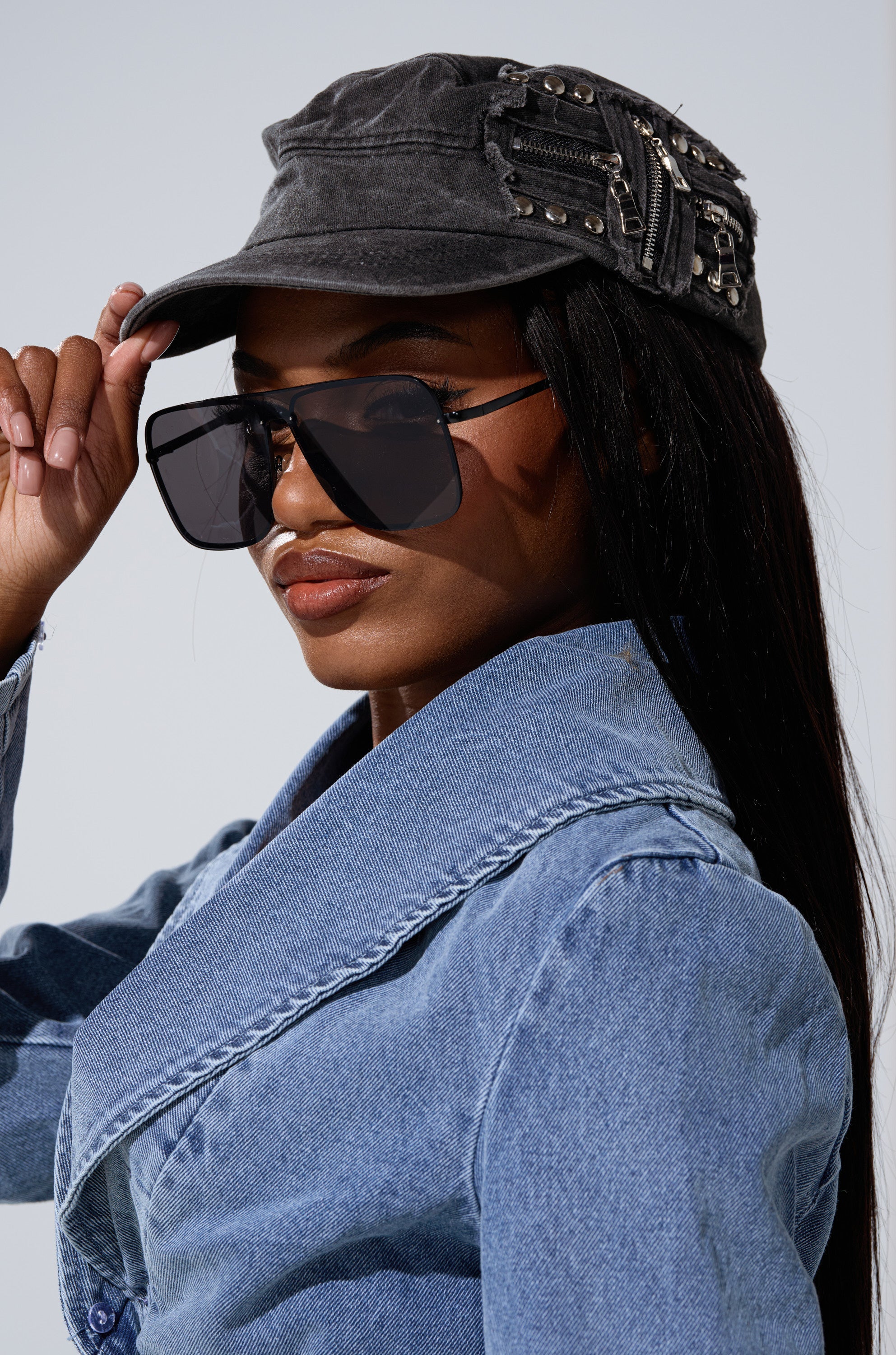 A woman adjusts the "I REALLY DO THIS HAT," a studded gray punk-rock cap, while wearing large black sunglasses and a light blue denim jacket, looking confidently at the camera. 0