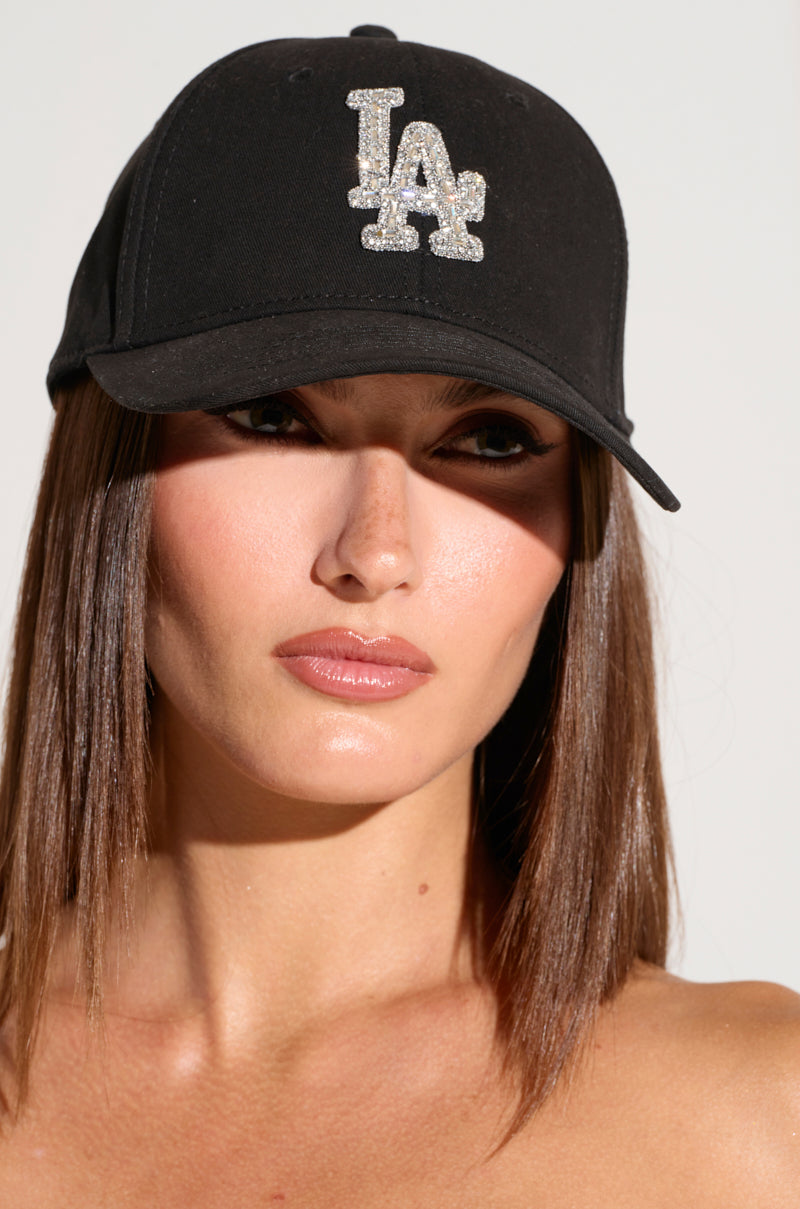 A person with straight, shoulder-length brown hair is wearing an IN THE TIMES SNAPBACK featuring a black design, an adjustable buckle, and the letters "LA" adorned with sparkly silver rhinestones on the front. They have a serious expression and are posed against a neutral background.