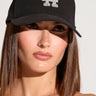 A person with straight, shoulder-length brown hair is wearing an IN THE TIMES SNAPBACK featuring a black design, an adjustable buckle, and the letters "LA" adorned with sparkly silver rhinestones on the front. They have a serious expression and are posed against a neutral background.