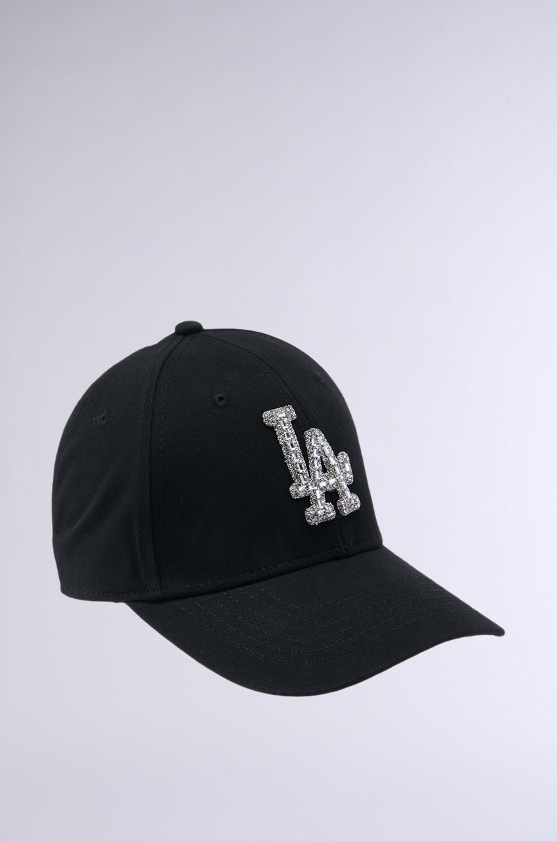 The IN THE TIMES SNAPBACK is a black baseball cap featuring the "LA" logo in white, positioned slightly to the left, set against a plain white background. The cap has a curved brim, six ventilation eyelets on the crown, and an adjustable buckle for a custom fit.