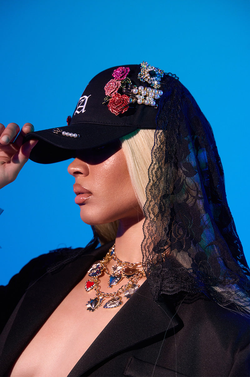 A person with long blonde hair wears the CITY GIRL LUXE BROOCH HAT— a black cap with floral and pearl details— plus a black lace veil, gold charm necklace, and black jacket, posing against a blue backdrop.