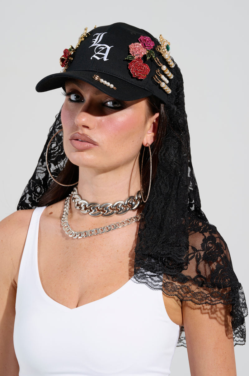 A woman in a white tank top is wearing the CITY GIRL LUXE BROOCH HAT, beautifully decorated with embroidery, gemstone brooches, and jewels. The hat features an adjustable buckled back strap and a lace veil. She accessorizes with large hoop earrings and chunky silver chain necklaces, maintaining a neutral expression complemented by dramatic makeup.