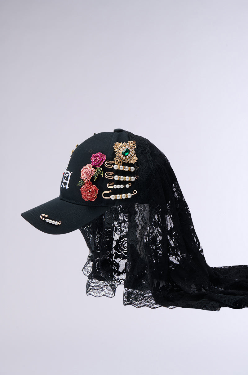 The CITY GIRL LUXE BROOCH HAT is a stylish black baseball cap adorned with vibrant floral embroidery, pearls, and decorative pins. It features an elegant drape of black lace fabric at the back and offers a perfect fit through its adjustable buckled back strap. The plain white backdrop accentuates its chic design.