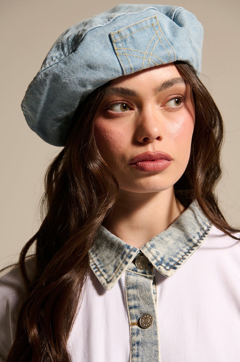A woman with long brown hair wears the IT GURL DENIM BERET by AKIRA Label and a white shirt with a light denim collar, gazing to the side with a neutral expression.