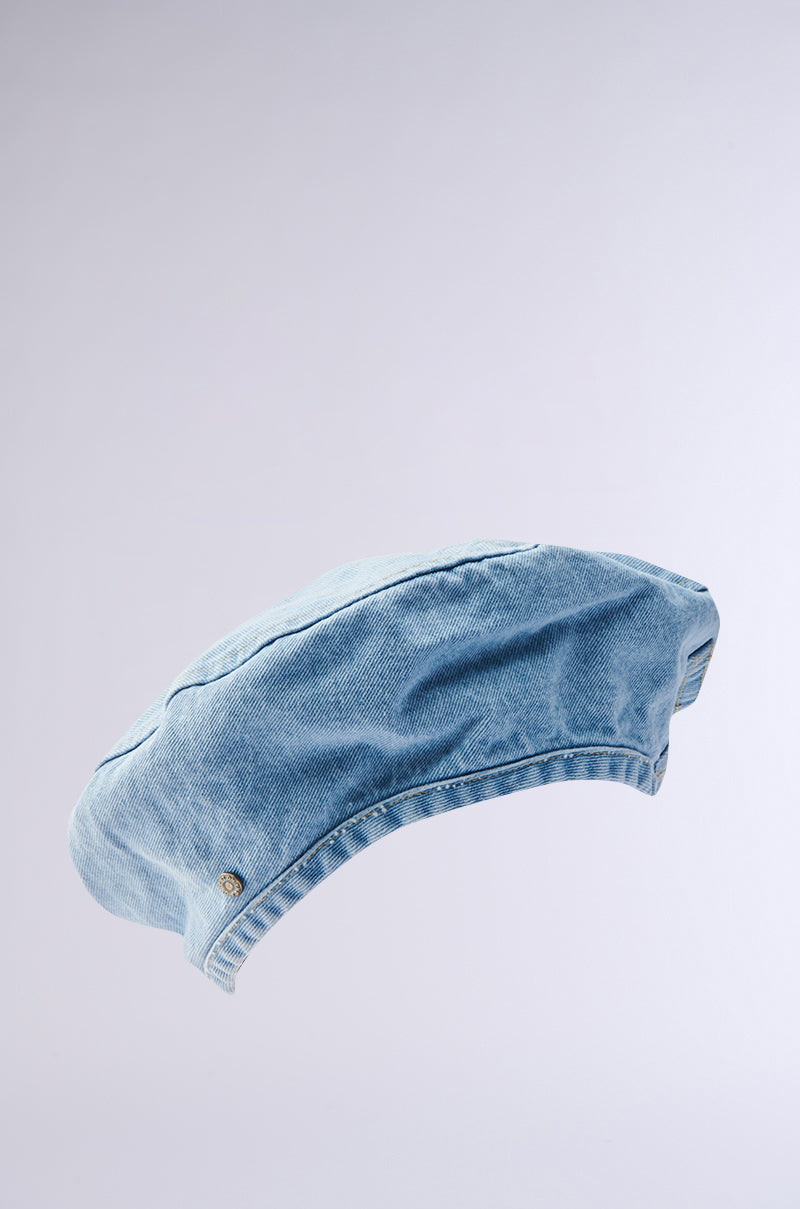 The IT GURL DENIM BERET from AKIRA Label, in light blue denim with a small metal button detail, is photographed on a plain light gray background—an effortlessly chic accessory for any outfit.