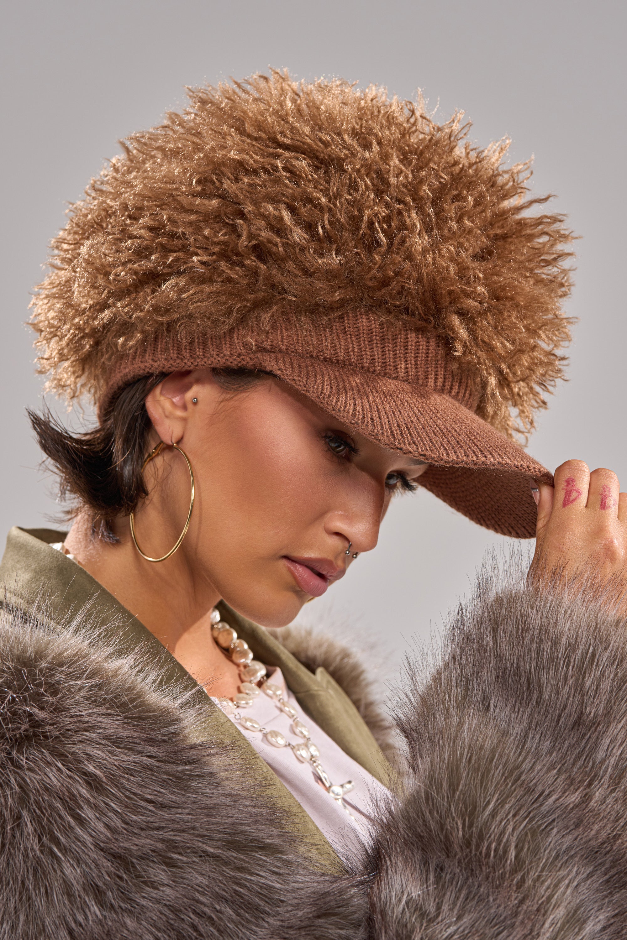 Wearing the WHOS HAIR HAT, a woman in a fleece-lined faux fur coat looks down, holding the brim. She accessorizes with hoop earrings, nose ring, and beaded necklace against a neutral gray background.