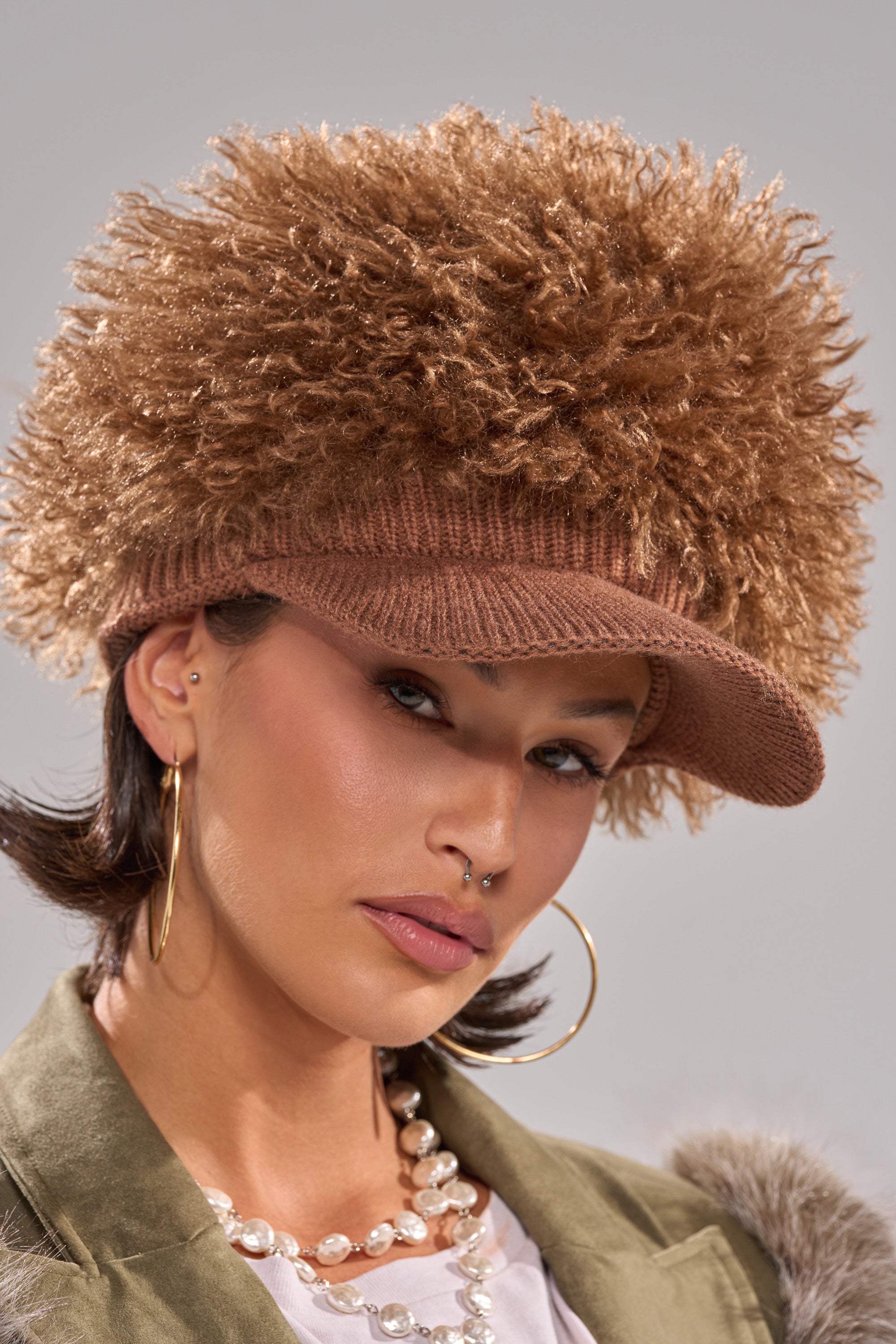 A woman wearing the WHOS HAIR HAT, featuring textured faux fur and a knitted brim, pairs it with large hoop earrings, a pearl necklace, and a nose ring, posing against a neutral background. 1