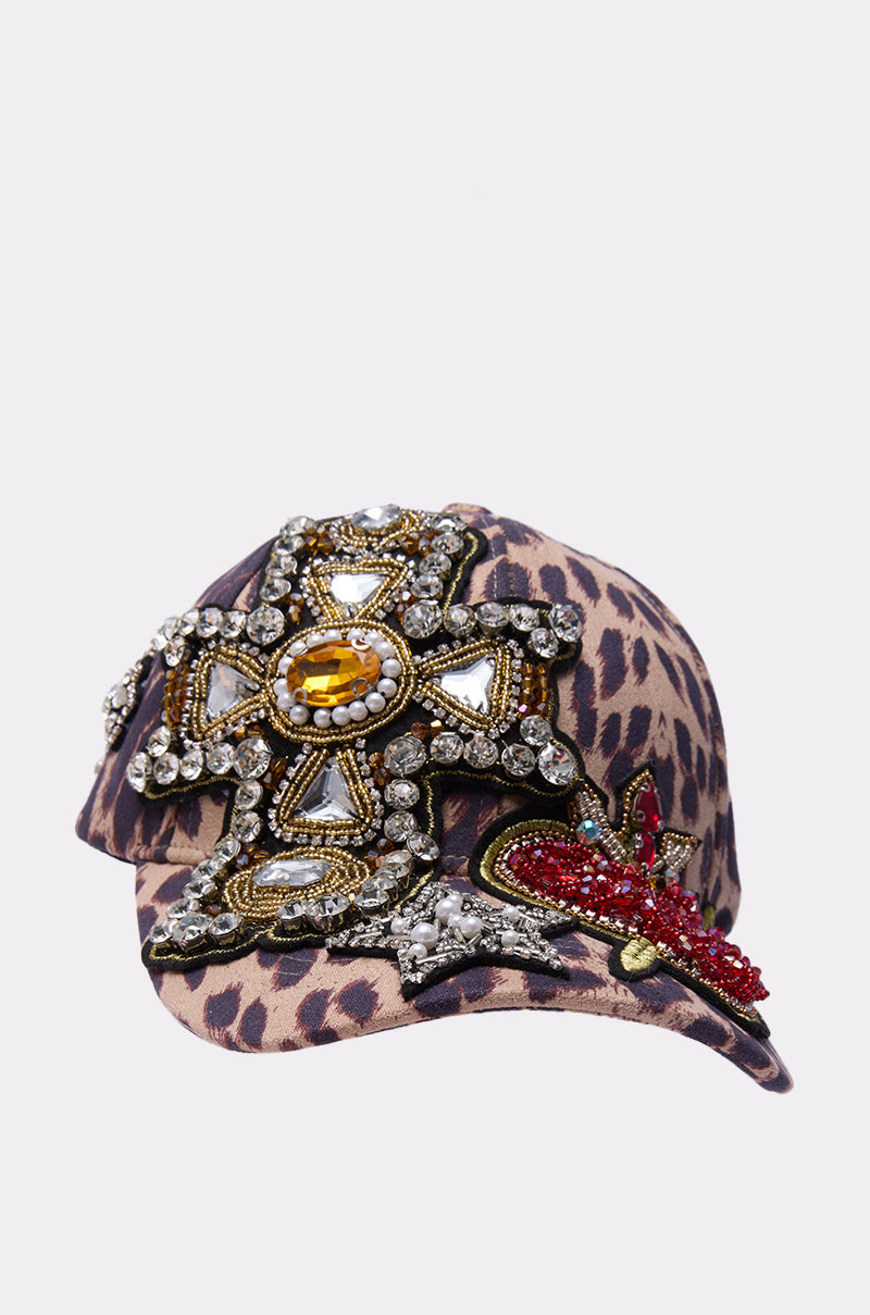 The GET ME TO MILAN PATCH HAT is a leopard-print baseball cap with a jeweled cross patch, decorated with pearls, gems, and a bold red beaded accent on the brim. 3