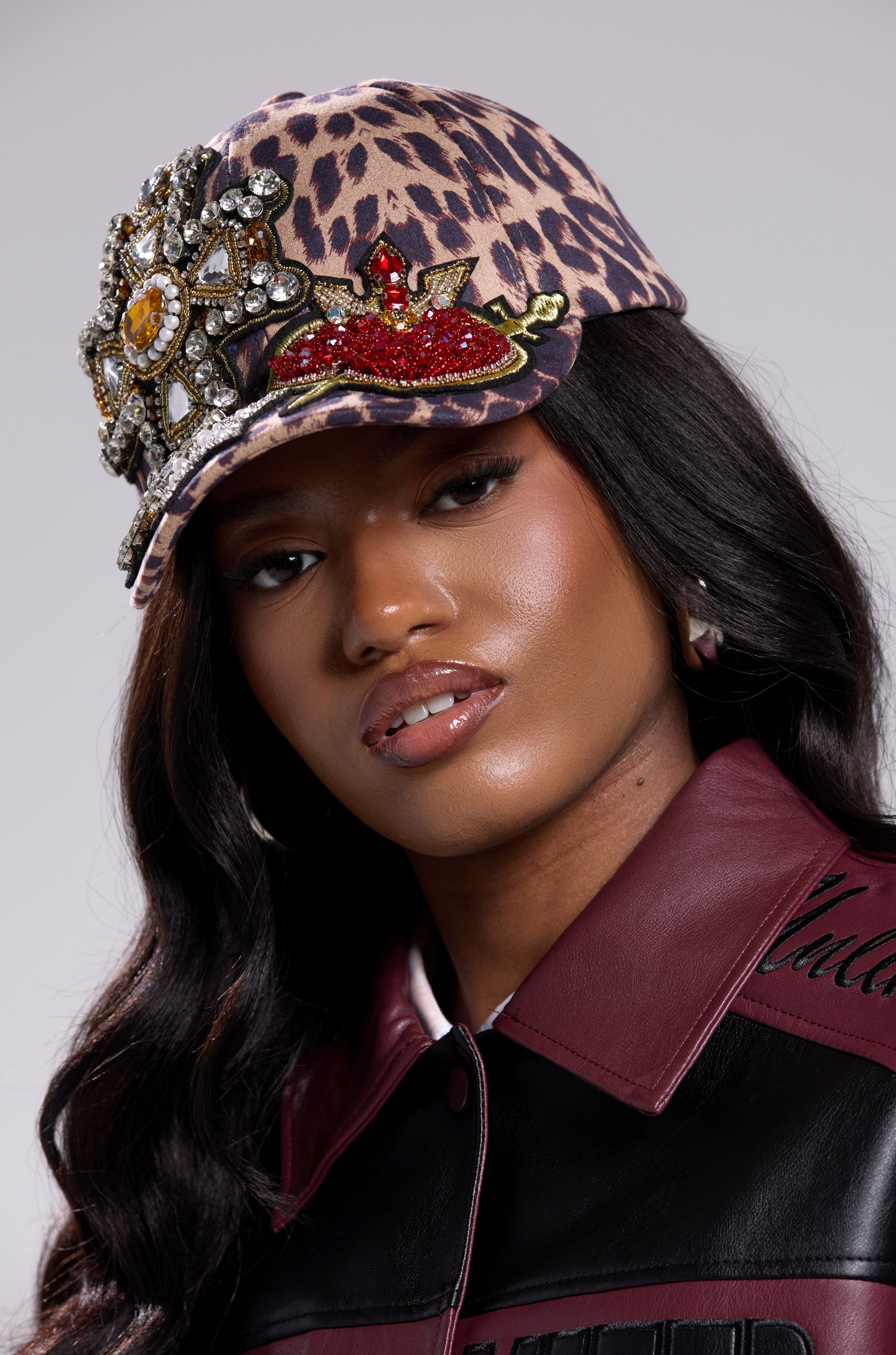 A woman with long wavy dark hair wears the GET ME TO MILAN PATCH HAT, a leopard-print baseball cap with jeweled patches and an adjustable back strap, paired with a black and burgundy leather jacket, looking confidently at the camera. 1