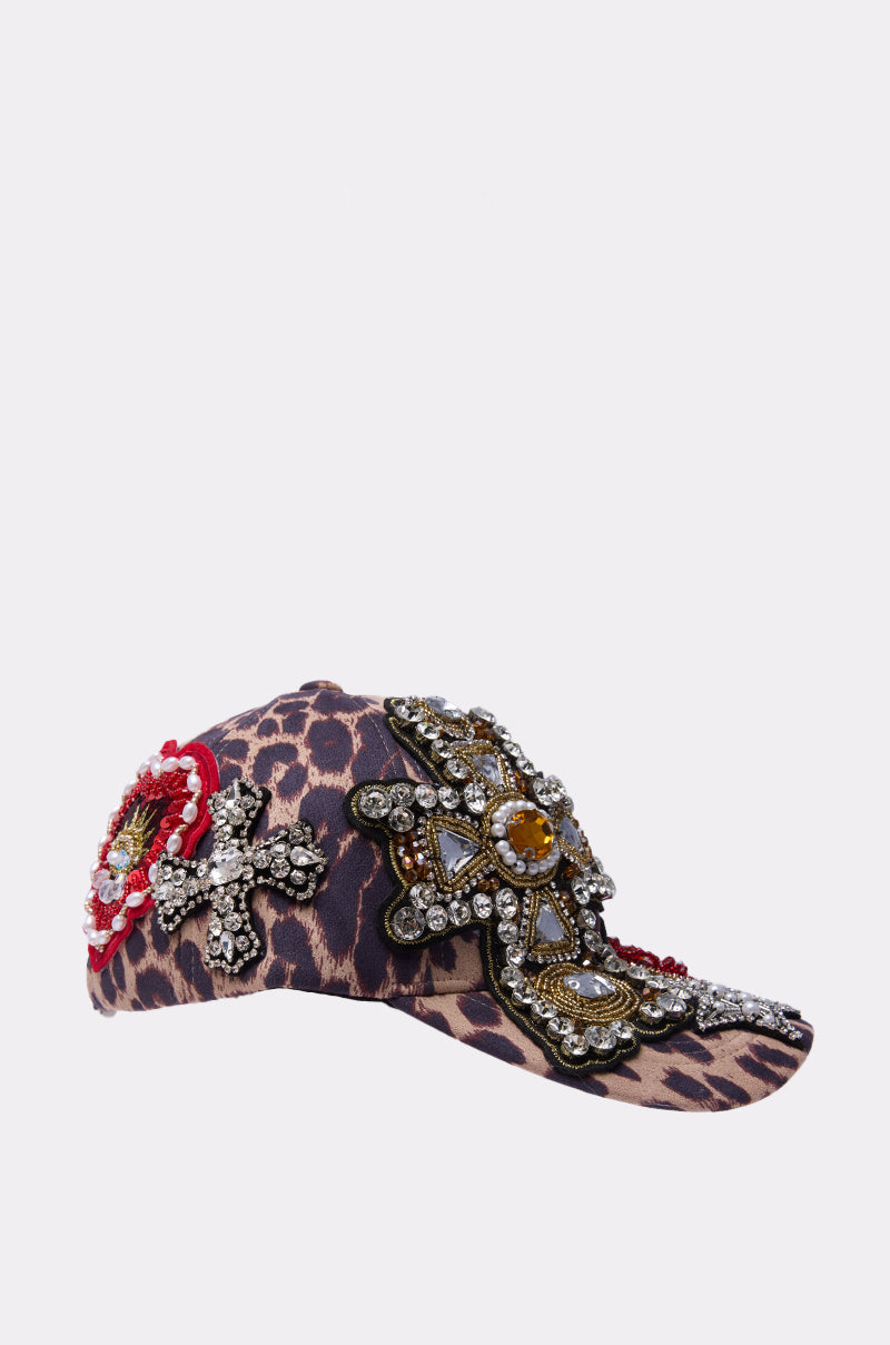 The GET ME TO MILAN PATCH HAT, a leopard-print baseball cap featuring ornate beads, jeweled embellishments, a large jeweled cross patch, and a floral motif, is displayed on a plain white background. 4