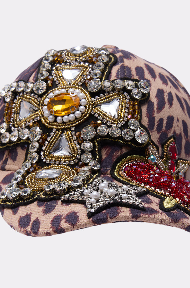 The GET ME TO MILAN PATCH HAT is a leopard-print baseball cap featuring an ornate jeweled cross patch with gold, pearl, and crystal accents, plus a red and silver beaded brim design. 5