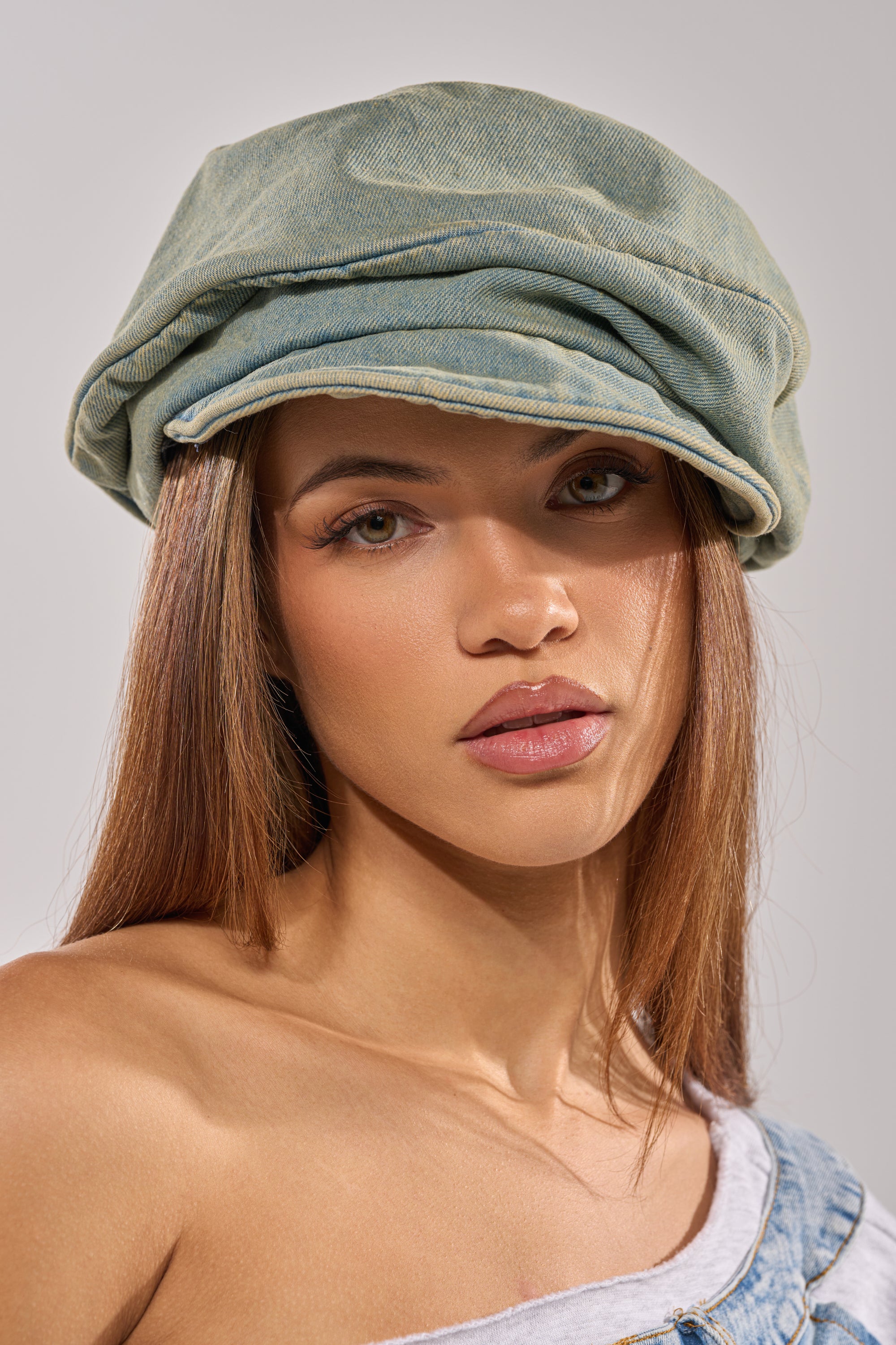 A woman with straight brown hair wears the IT GURL SLOUCH DENIM HAT in light green and an off-shoulder denim top, channeling effortless streetwear style as she gazes neutrally at the camera against a plain background.