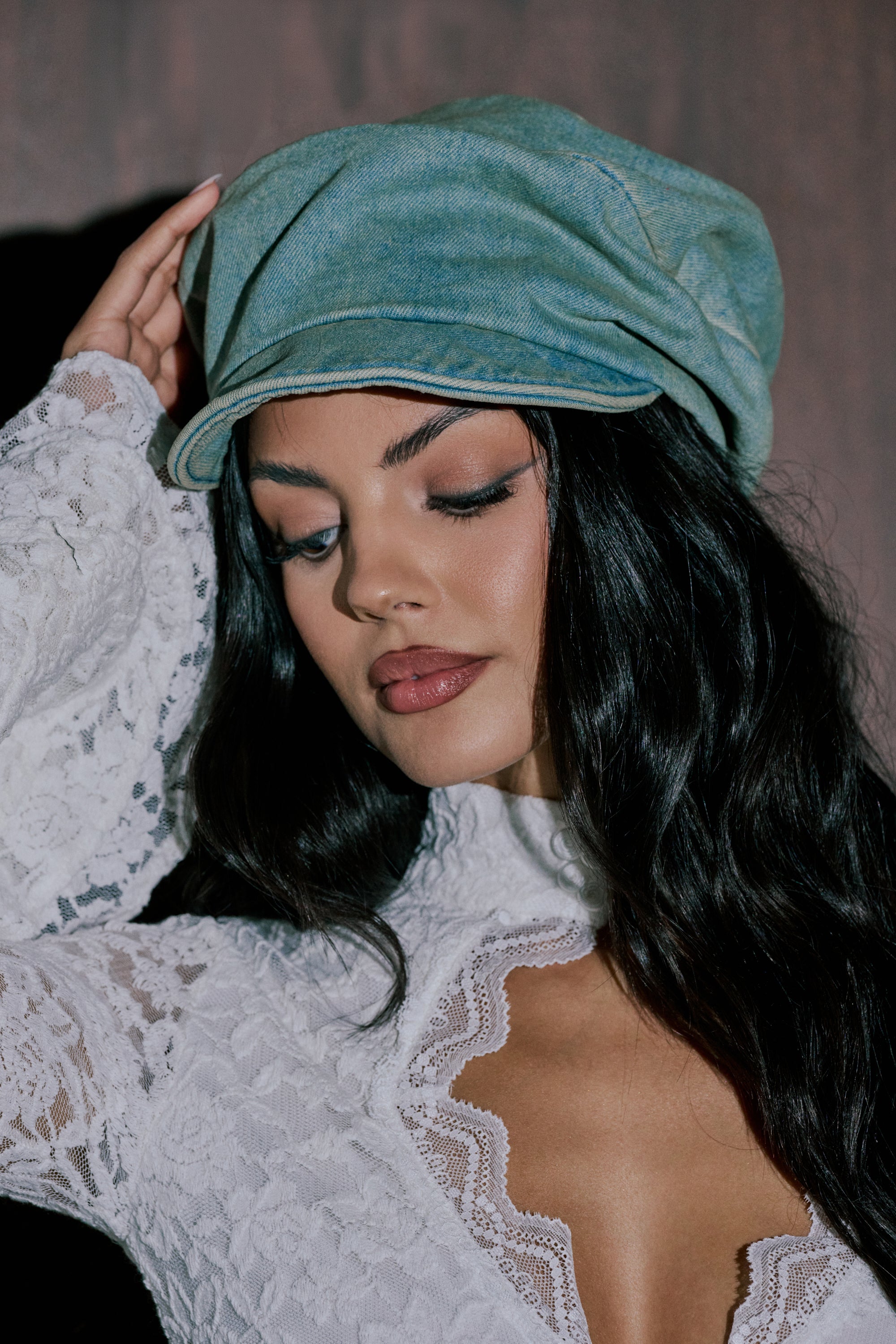 A woman with long dark hair wears the IT GURL SLOUCH DENIM HAT and a white lace top, looking down as she touches the hat. The scene features a softly blurred background. 0