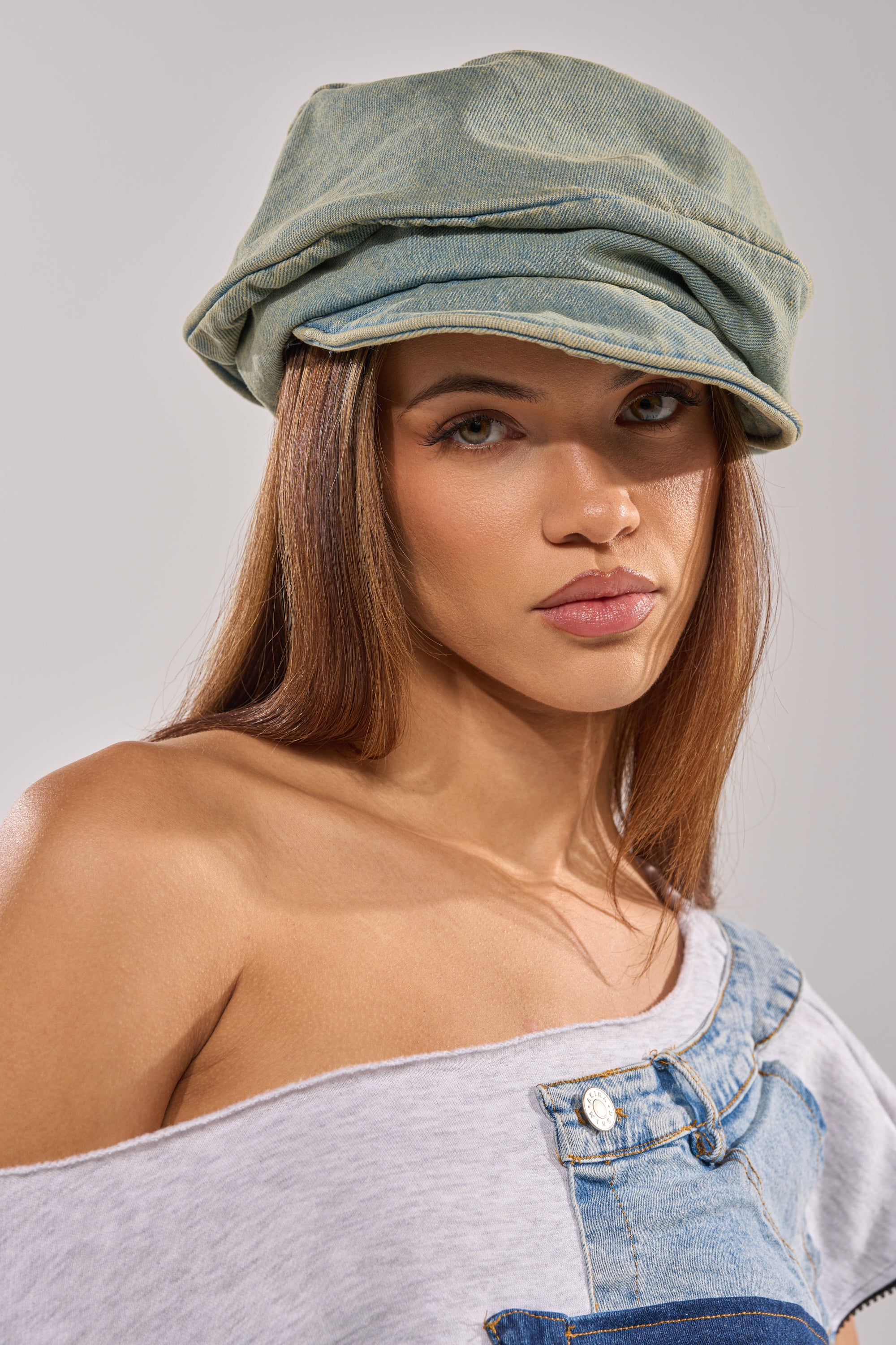 A woman with straight brown hair wears the IT GURL SLOUCH DENIM HAT and a casual off-the-shoulder gray and denim top, looking directly at the camera with a neutral expression.
