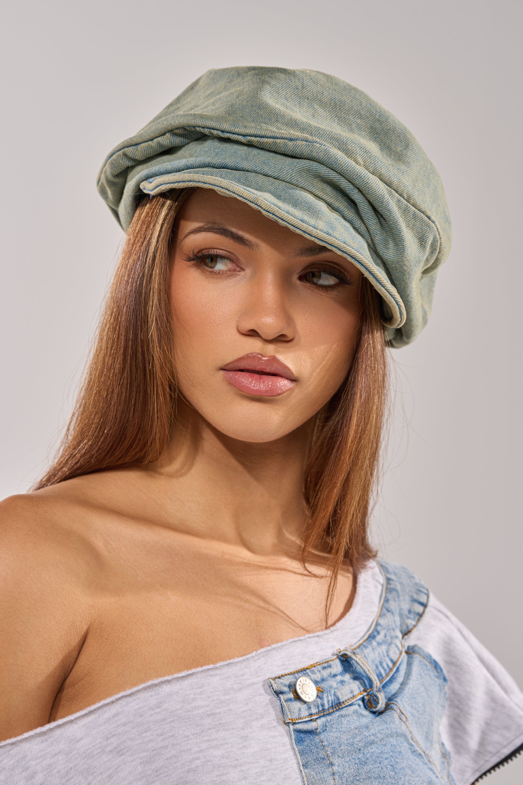 A young woman with long brown hair wears the IT GURL SLOUCH DENIM HAT, a gray off-the-shoulder top, and denim overalls, looking slightly to the side with a neutral expression.