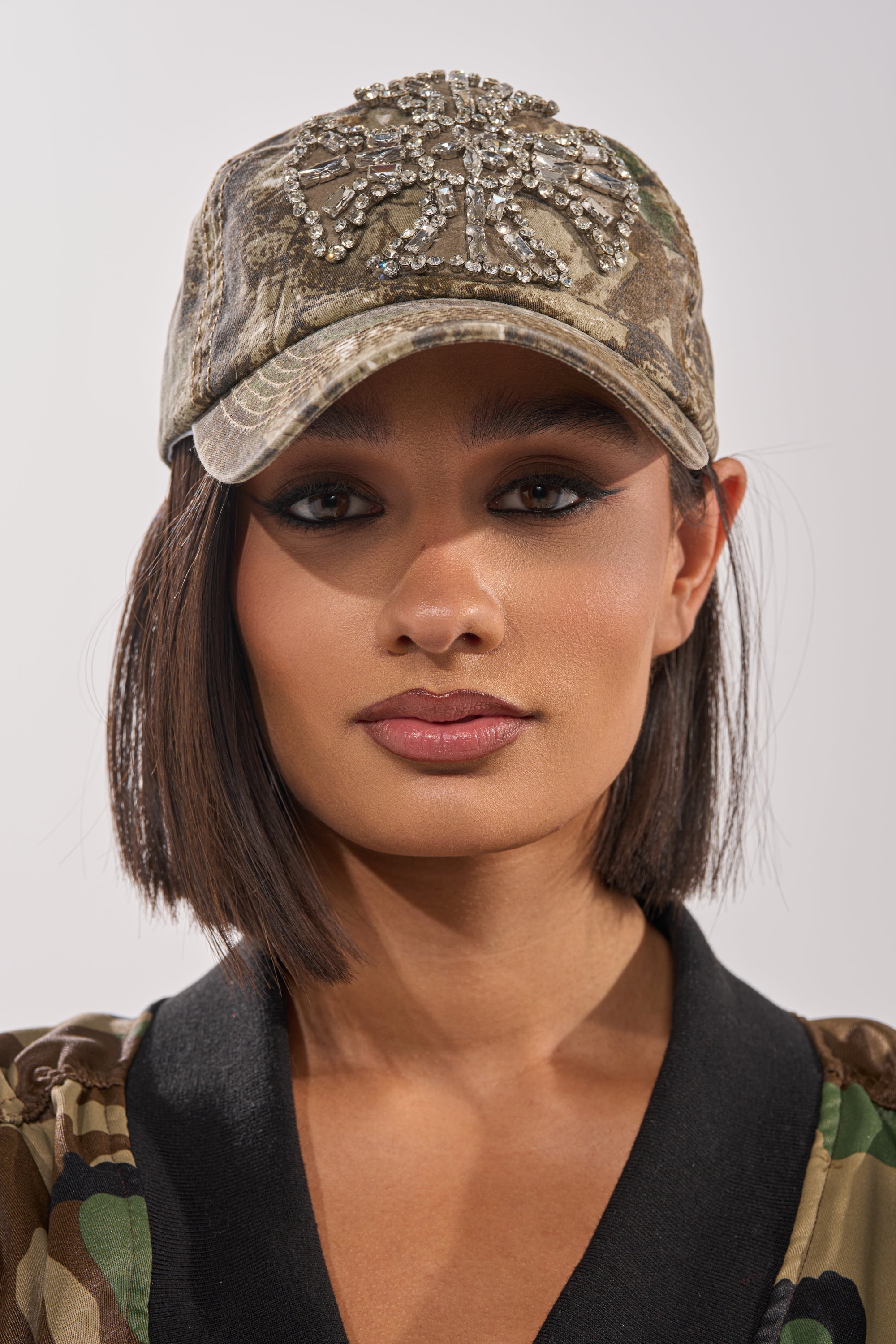 A woman with straight dark hair wears the SEE ME APPLIQUE HAT and a matching camo jacket, looking directly at the camera with a neutral expression. The sparkling hat adds flair to her bold street-style look. 2