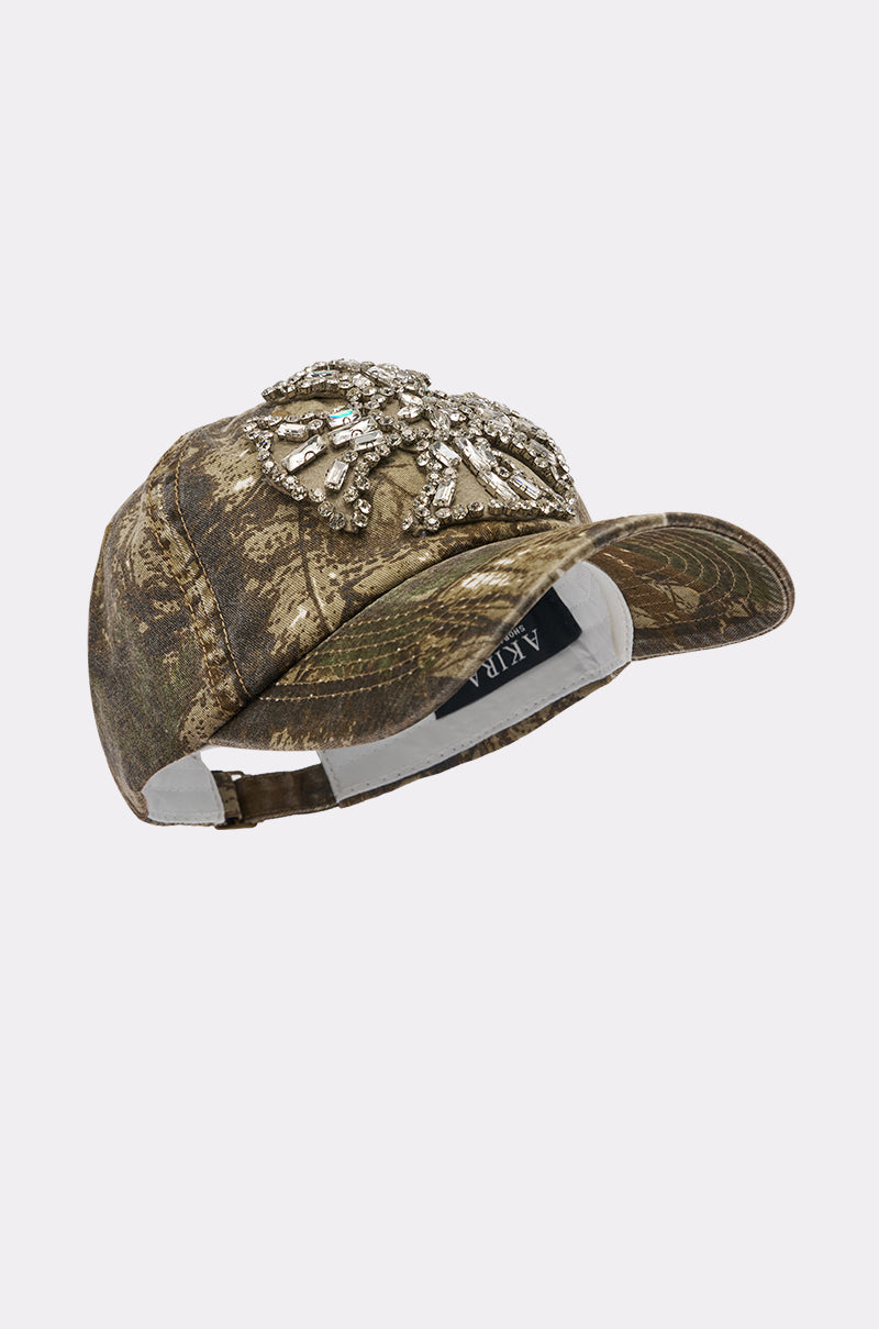 The SEE ME APPLIQUE HAT is a camo baseball cap adorned with a cluster of silver beads and crystals on the front, making it a standout street-style accessory. Displayed against a simple white background. 4
