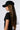 A person with long, straight brown hair is wearing the PROUD OF ME RHINESTONE SNAPBACK, which is a black baseball cap with a fishnet mesh veil extending from the back. They are also sporting a fitted black T-shirt. The background is plain and light-colored.