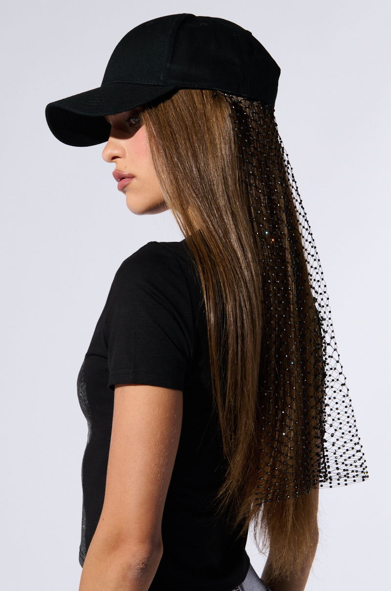 A person with long, straight brown hair is wearing the PROUD OF ME RHINESTONE SNAPBACK, which is a black baseball cap with a fishnet mesh veil extending from the back. They are also sporting a fitted black T-shirt. The background is plain and light-colored. 0