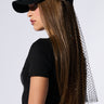 A person with long, straight brown hair is wearing the PROUD OF ME RHINESTONE SNAPBACK, which is a black baseball cap with a fishnet mesh veil extending from the back. They are also sporting a fitted black T-shirt. The background is plain and light-colored.