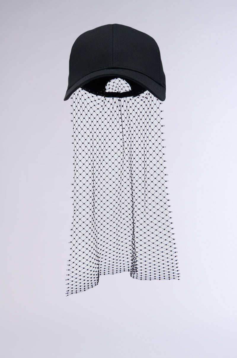 The PROUD OF ME RHINESTONE SNAPBACK is a black baseball cap with an adjustable strap, paired with a hanging, net-like veil made of white fabric that features a grid pattern of small black dots. The veil extends downward to cover where the face would be when worn. The background is plain light gray. 2