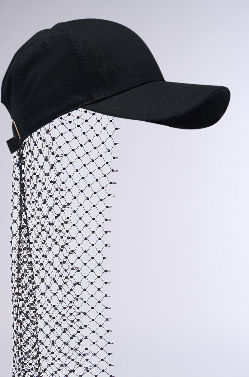 The PROUD OF ME RHINESTONE SNAPBACK is a black baseball cap featuring an attached black fishnet mesh veil that drapes over the back and extends downward. The veil showcases a diamond-patterned design formed by thin, intersecting lines. The background of the product is plain light gray, and the cap includes an adjustable strap to ensure a perfect fit. 4