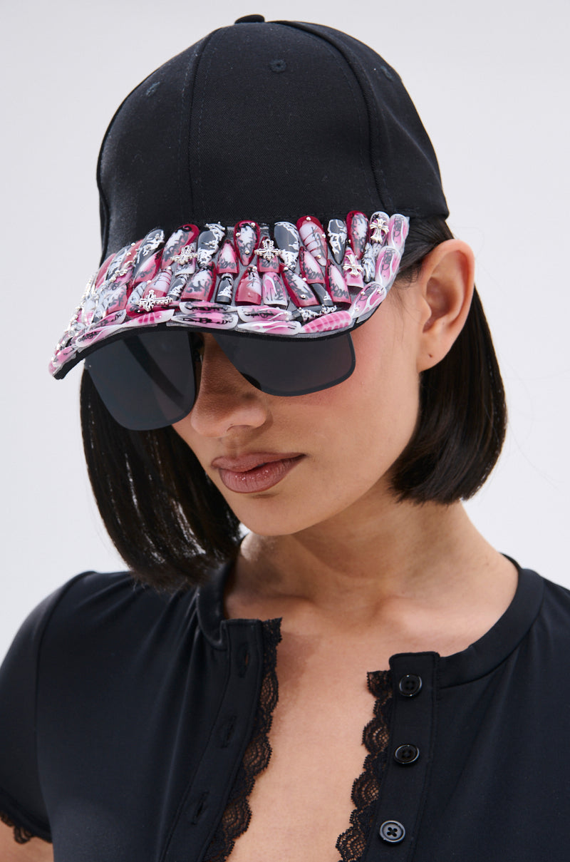 A woman wears the NAILED IT HAT, a black ball cap decorated with pink and black press-on nail accents on the brim, styled with oversized sunglasses and a black lace-trim buttoned top.