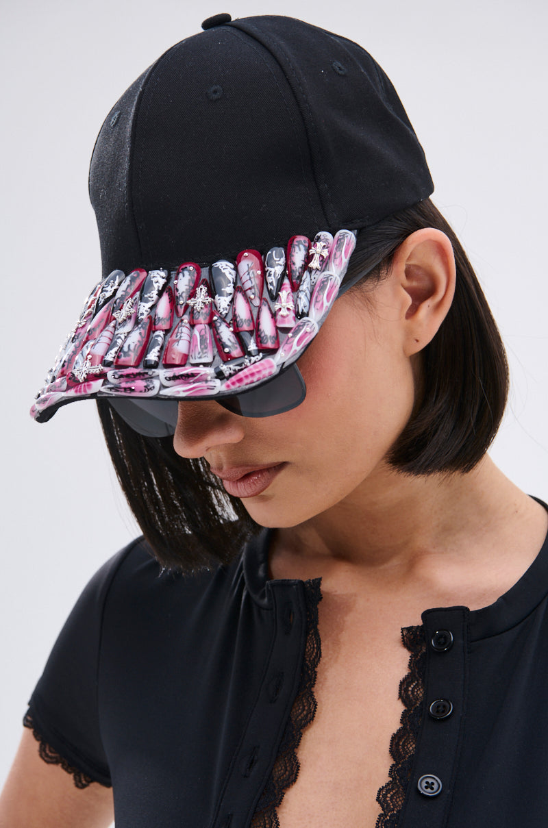 A woman in a bob haircut, lace-trimmed top, and sunglasses wears the NAILED IT HAT—a black ball cap featuring press-on nail embellishments on the brim—as she faces slightly downward in style.