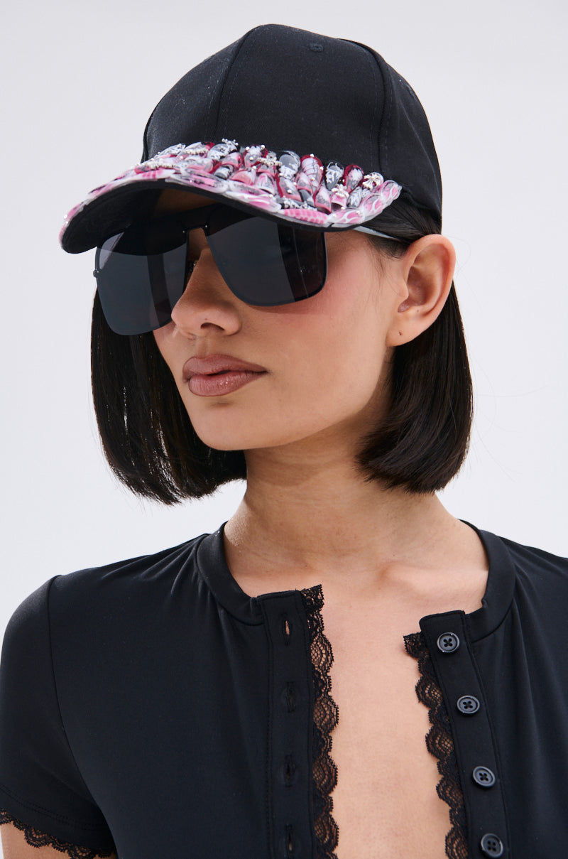 A woman with a black bob wears the NAILED IT HAT, featuring pink and white trim, along with oversized black sunglasses and a black lace-trimmed top. She faces forward against a plain light background.